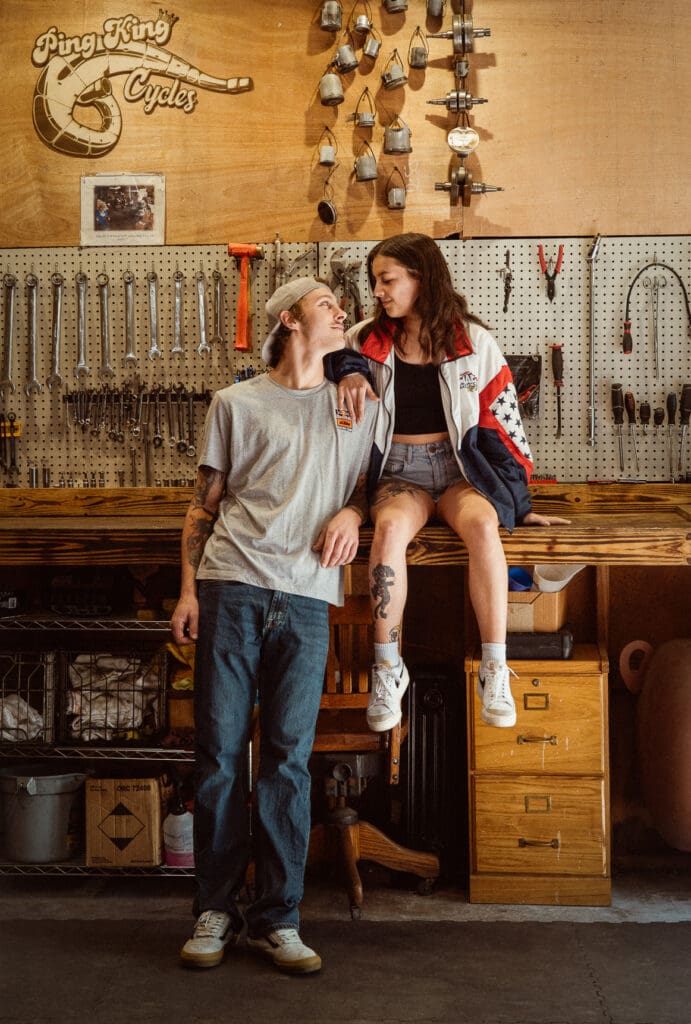 Editorial couple sitting in workshop setting with casual layered outfits, relaxed, vintage styling for creative engagement photo outfit inspiration. 