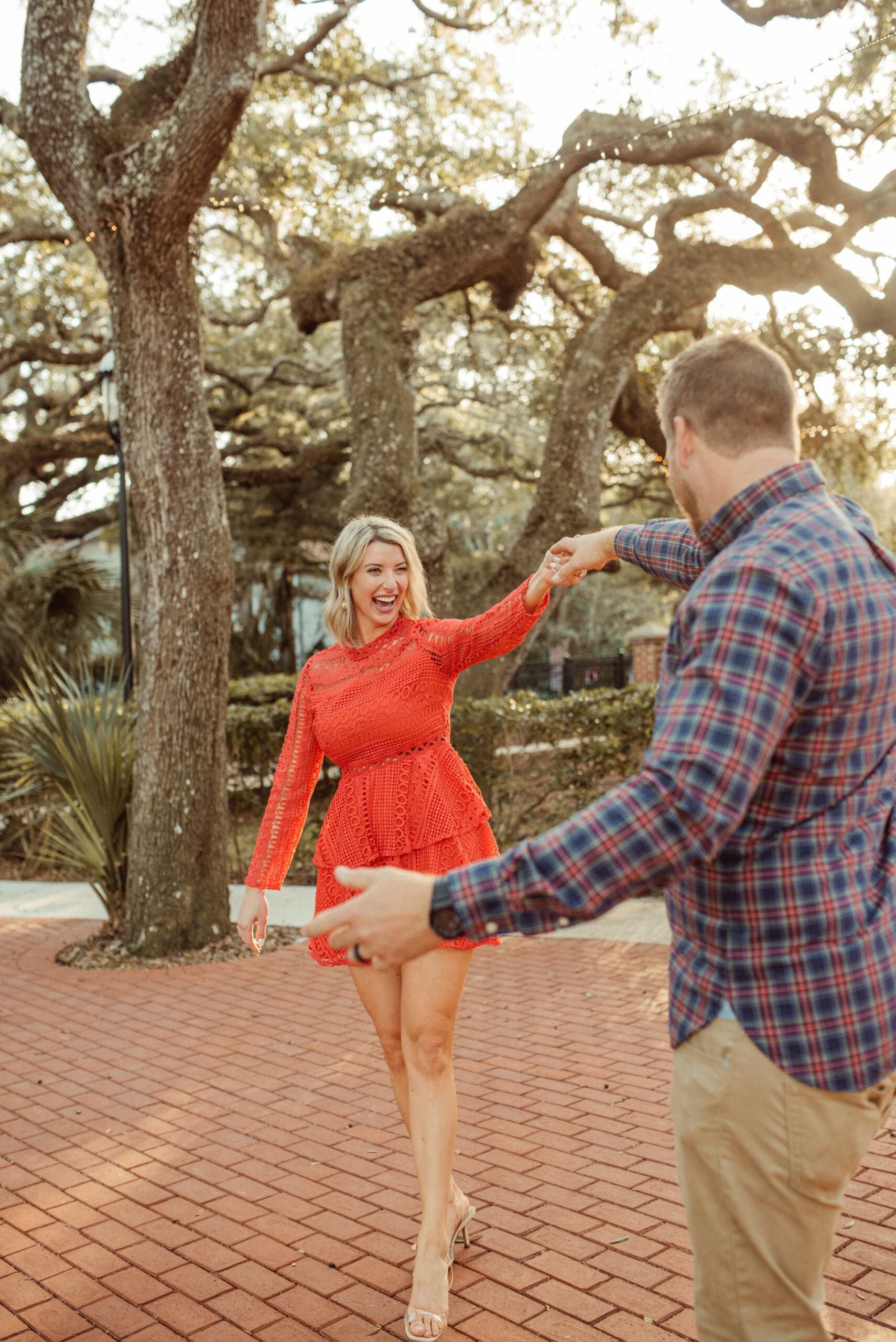 Couple spinning together under oak trees with a red dress adding movement and personality to a romantic engagement session. 