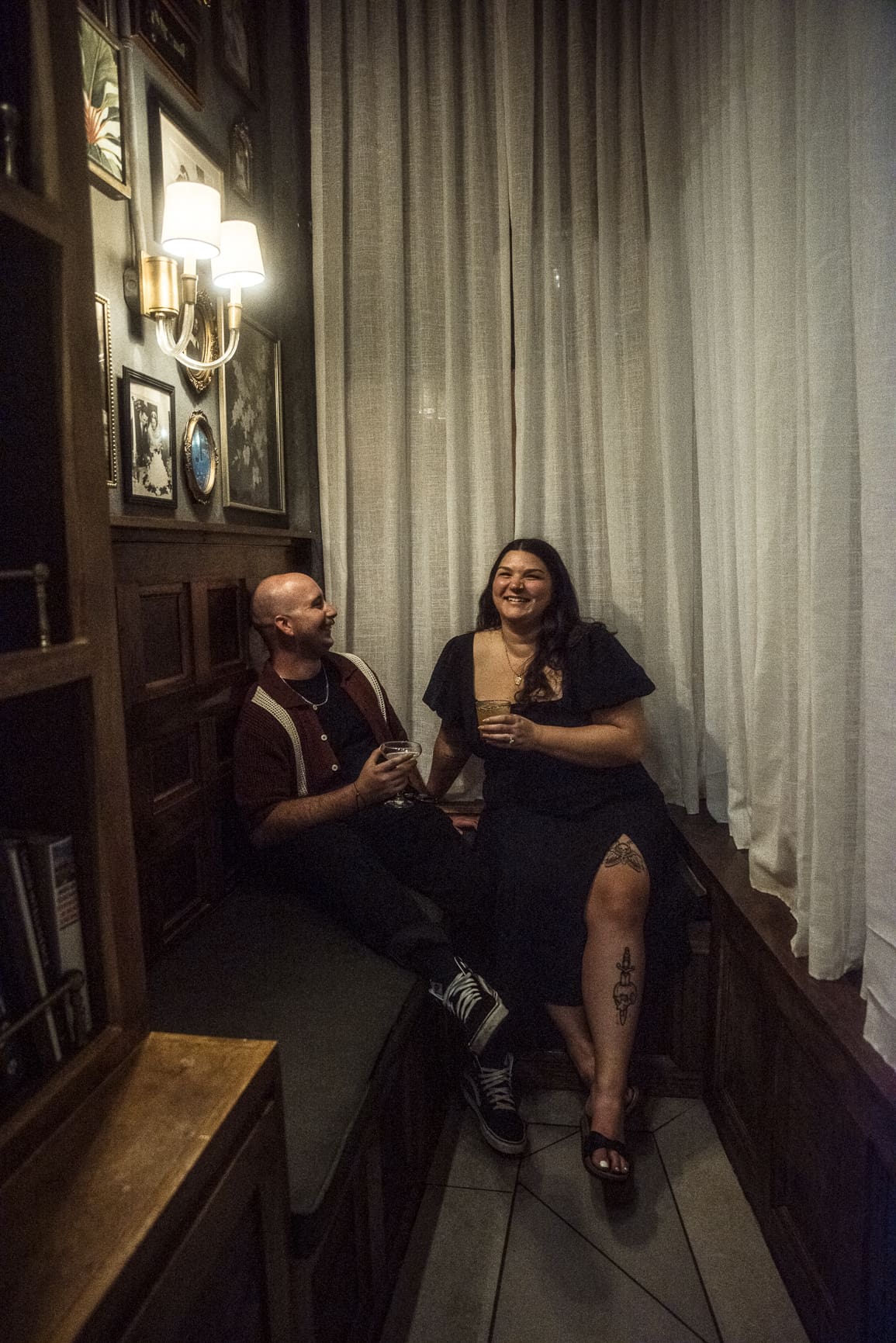 Couple sitting together indoors sharing drinks and laughing, cozy playful engagement session with natural interaction. 