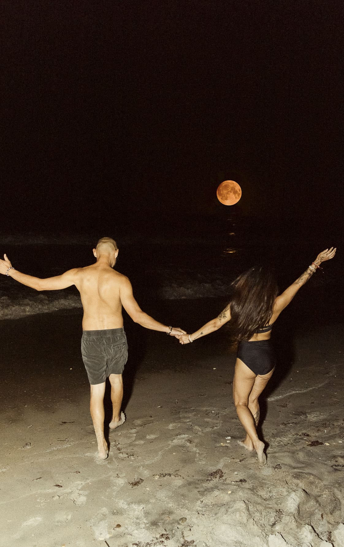 Couple running hand in hand on the beach at night with a glowing full moon behind them