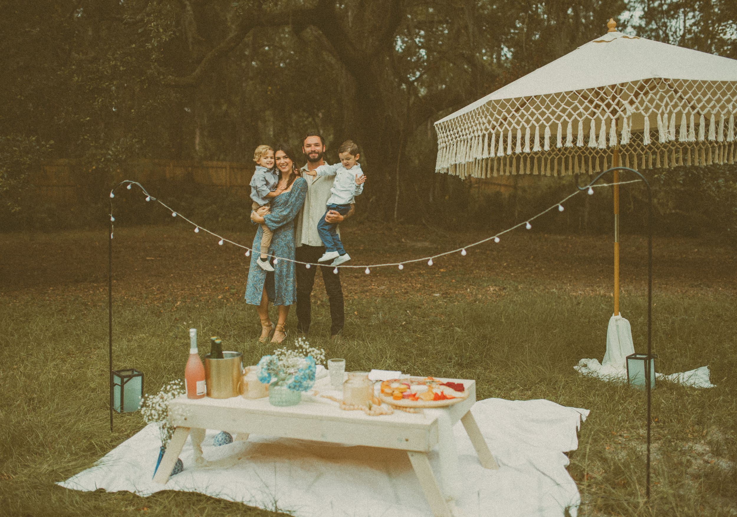 Couple holding their children near a styled picnic setup during a backyard engagement session in Jacksonville, Florida.