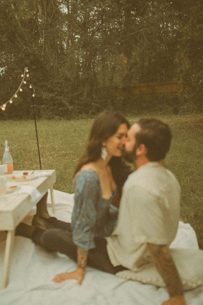Couple kissing on blanket during backyard picnic engagement session in Jacksonville with soft film tones.