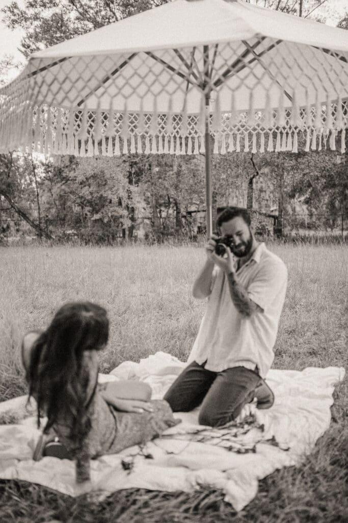 Groom taking photos with film camera during backyard engagement session black and white candid moment.