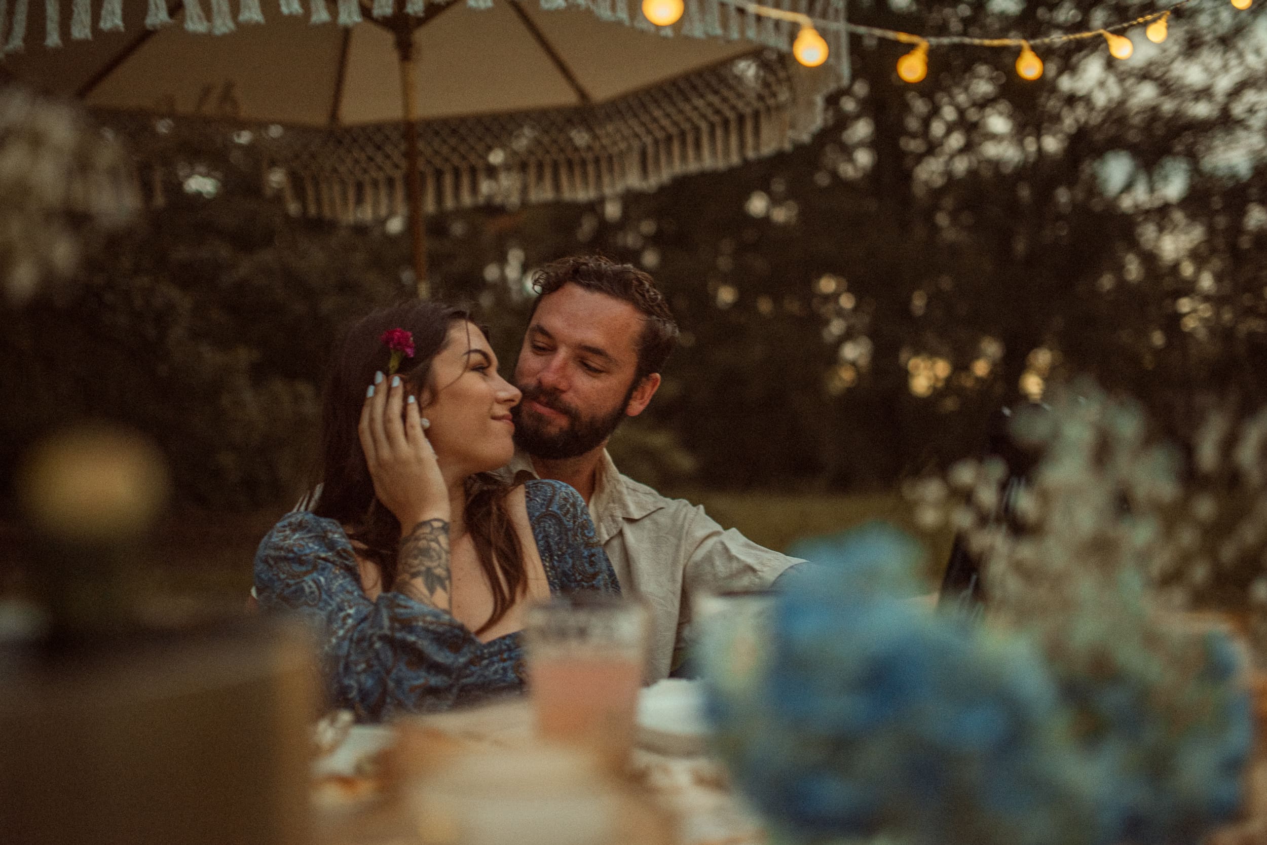 Couple sitting close together under string lights and a fringed umbrella during a backyard engagement session.