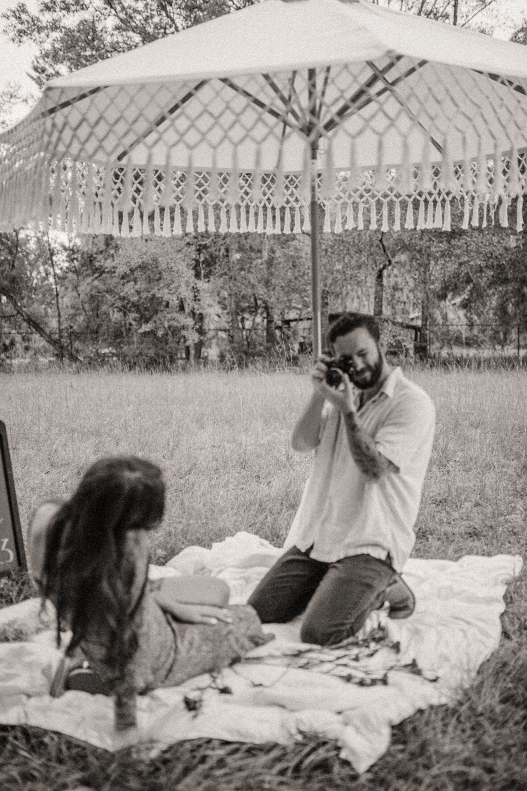 Black and white film photo of couple taking pictures of each other with vintage camera during an outdoor picnic under umbrella. 