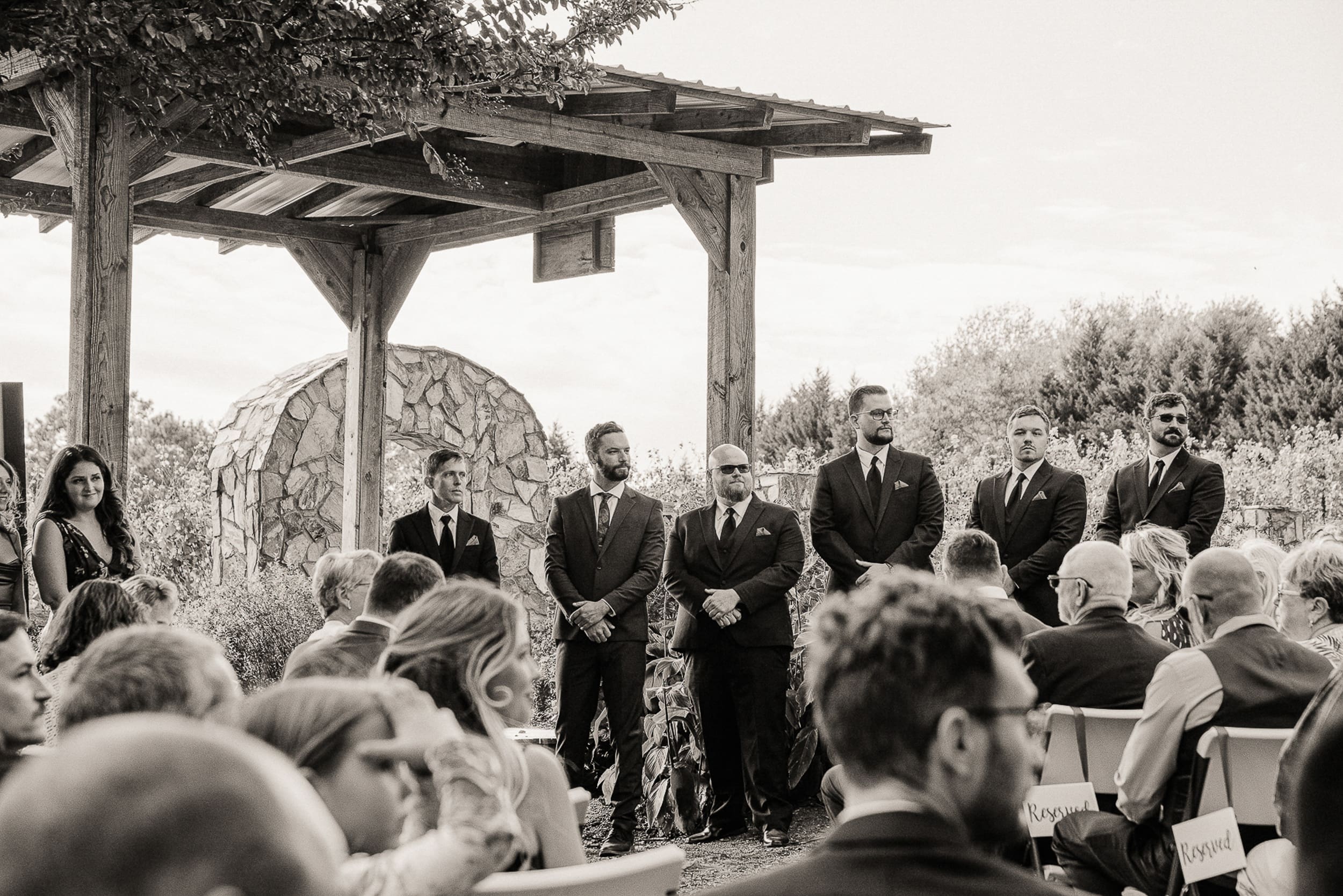 Black and white photo of groom and groomsmen waiting at outdoor altar under rustic pergola at Parker-Binna Vineyard North Carolina. 