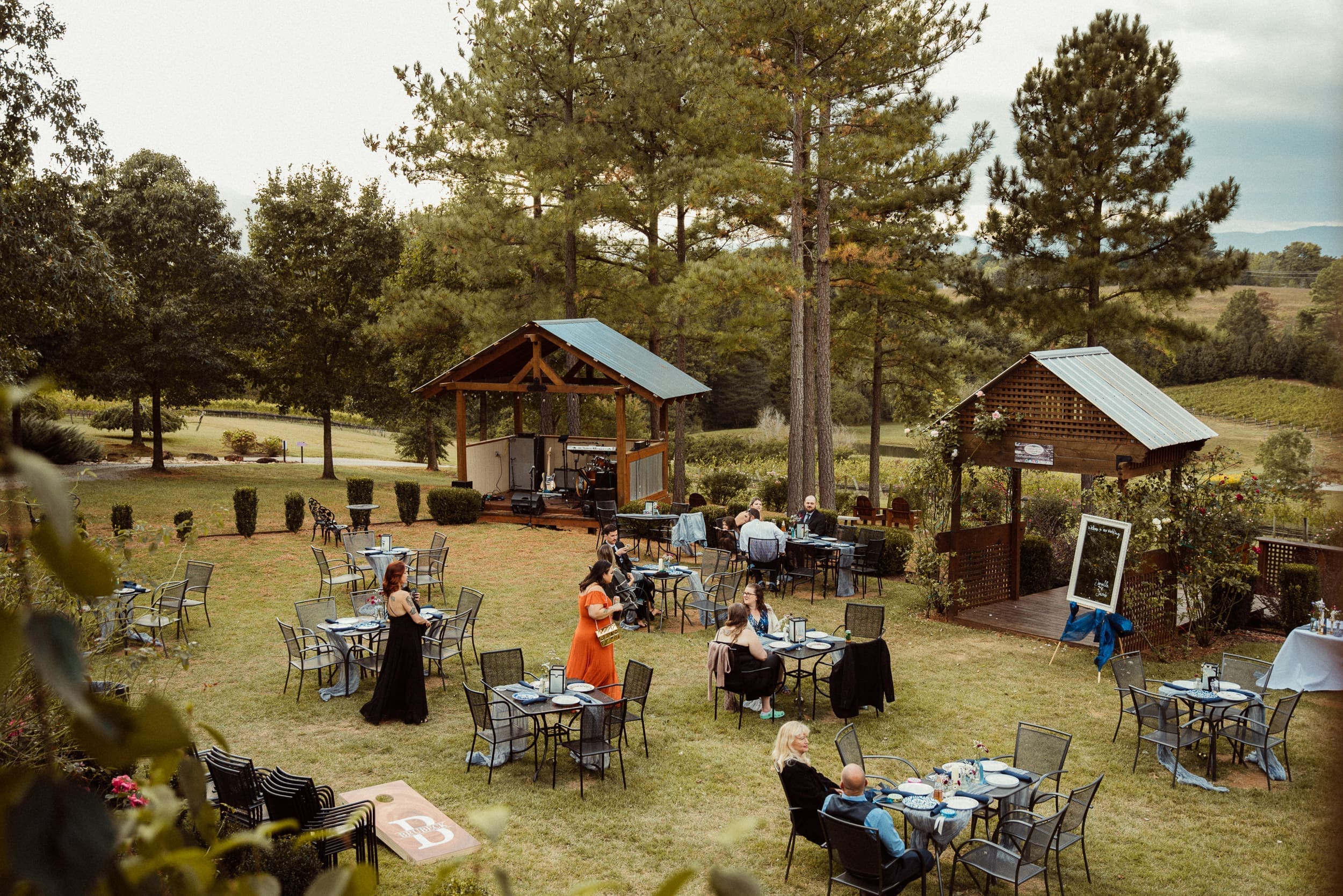 Wide overhead view of outdoor wedding reception with guests at tables and rustic pavilion stage at Parker-Binns Vineyard North Carolina. 