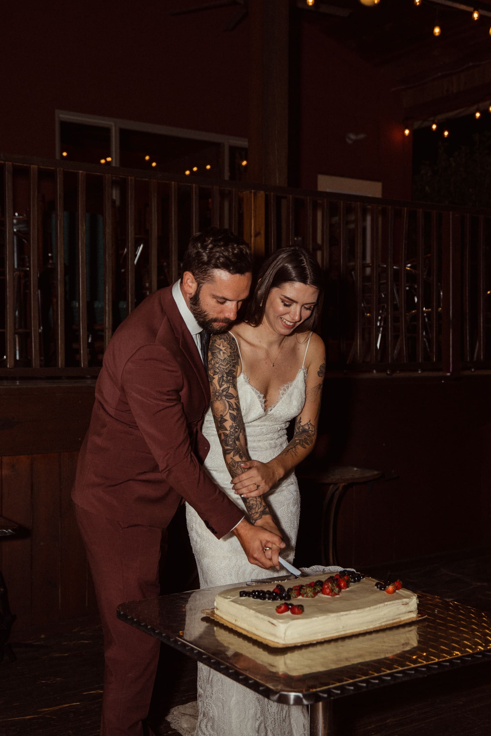 Bride and groom cutting wedding cake inside Parker-Binns Vineyard restaurant with string lights behind, North Carolina. 