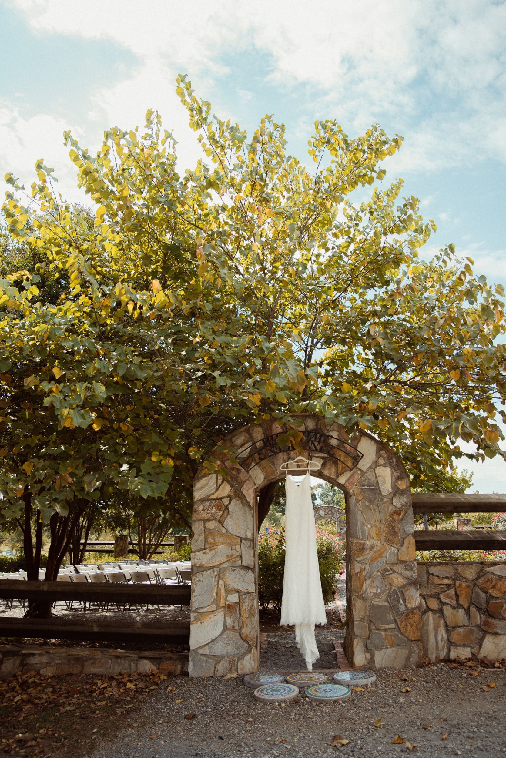 Wedding dress hanging in stone garden arch at Parker-Binns Vineyard with autumn vineyard and blue sky behind, North Carolina wedding photographer Brandi Denslow. 