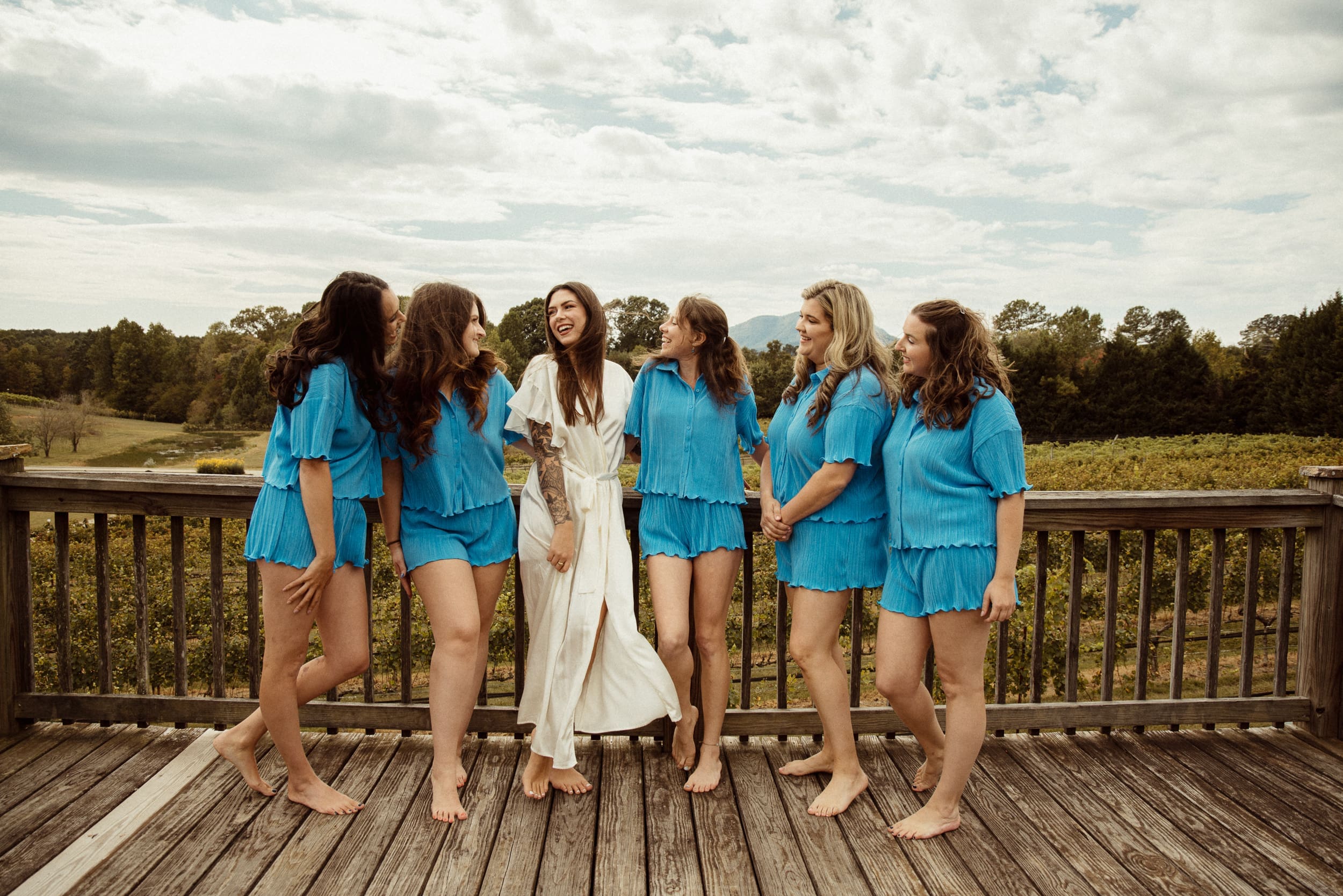Bride laughing with bridesmaids in matching blue sets on vineyard porch with Blue Ridge Mountains behind, Parker-Binns Vineyard North Carolina. 