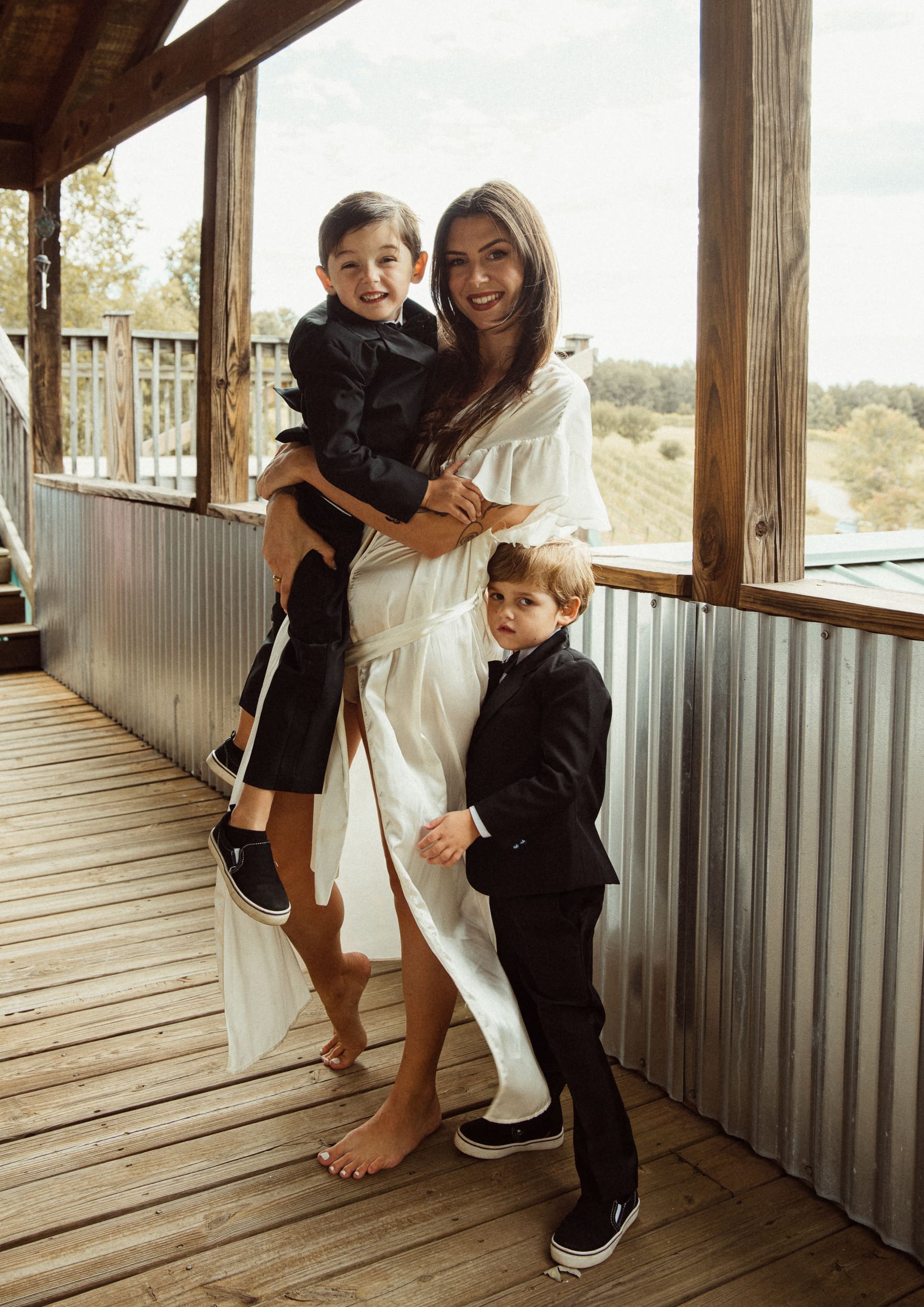 Bride in white robe holding her young son with second son beside her on rustic porch at Parker-Binns Vineyard, North Carolina wedding photography by Brandi Denslow. 