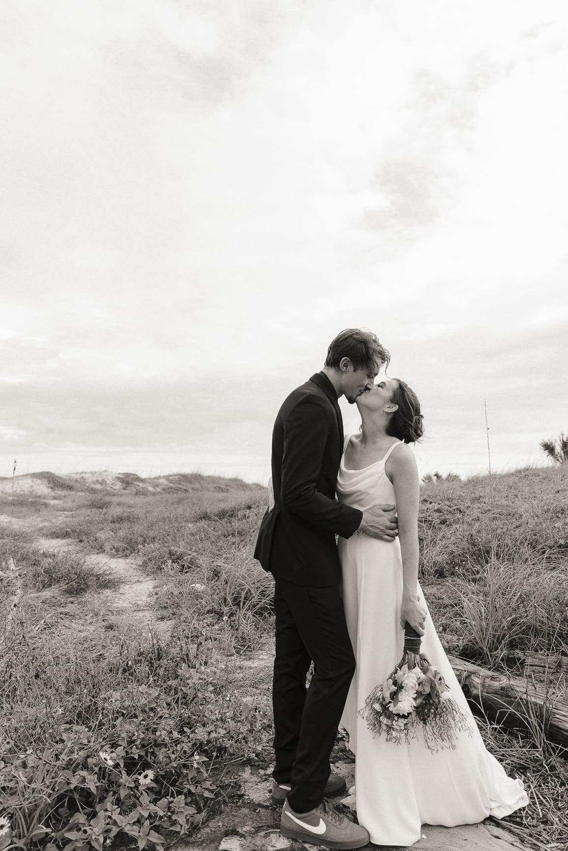 Bride and groom sharing first kiss on coastal dunes at sunset, film-toned wedding photography by Brandi Denslow St. Augustine. 