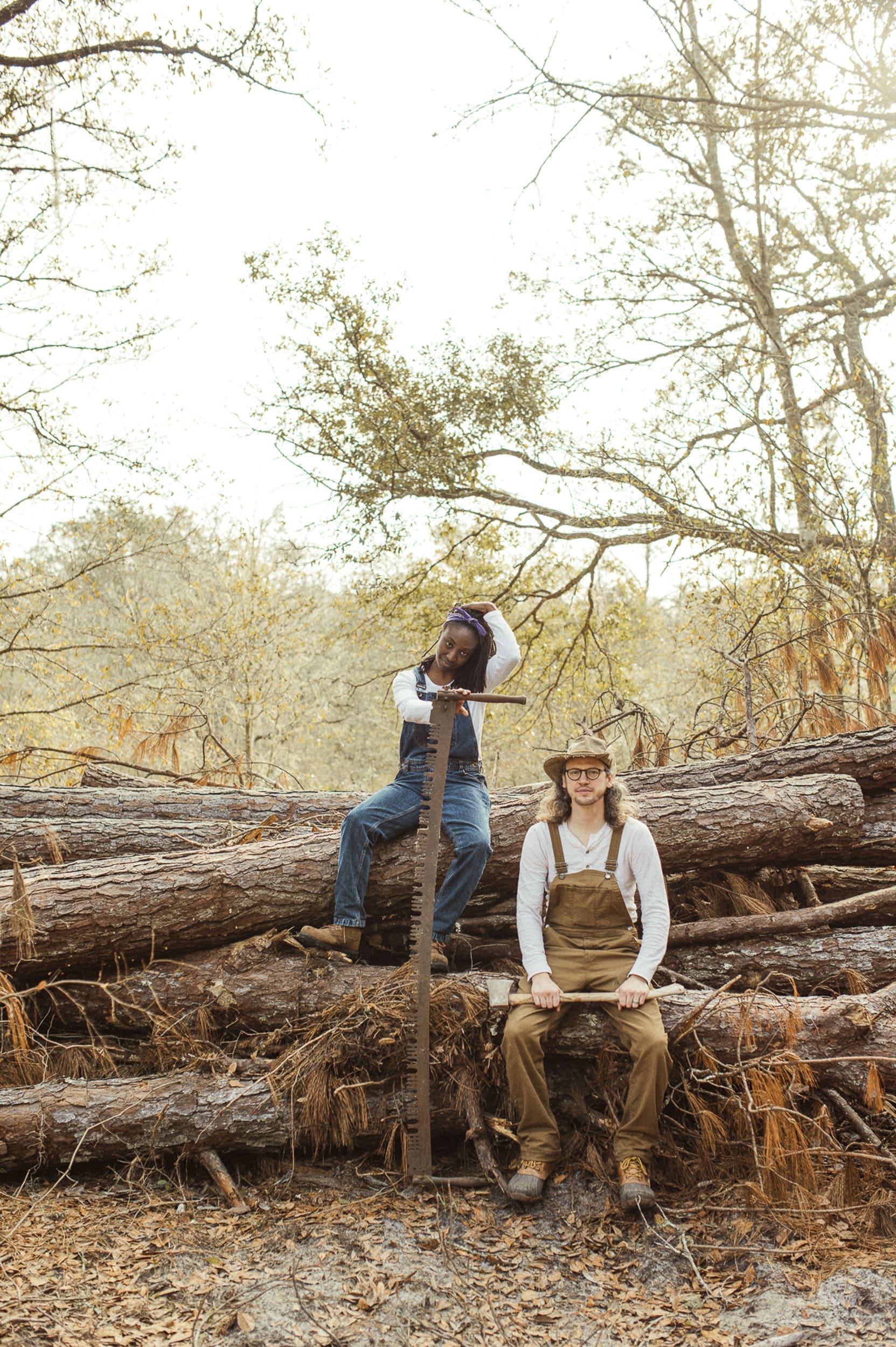 Editorial engagement portrait of a couple sitting on fallen logs with an antique saw during a Florida farm session. 