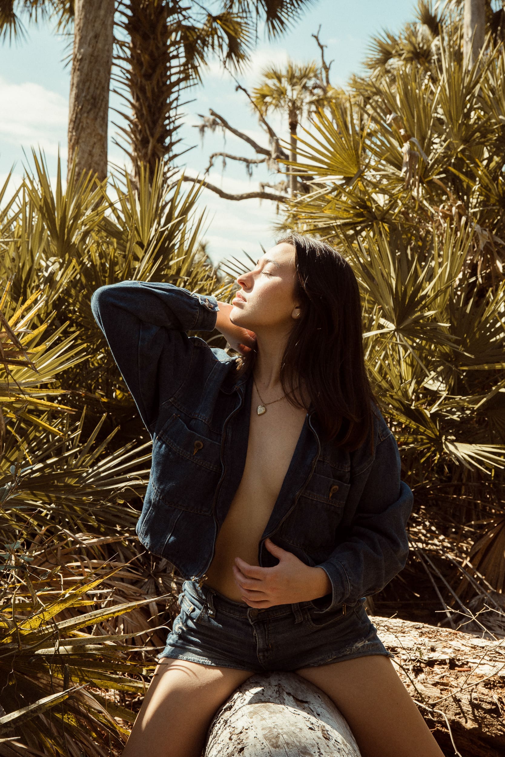 Woman standing in nature with an open denim jacket, eyes closed toward the sunlight during an editorial outdoor boudoir and empowerment portrait session in St. Augustine, Florida.