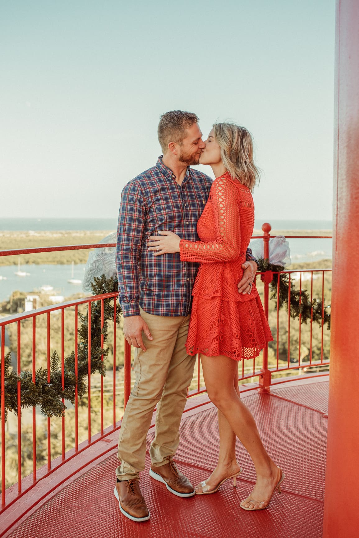 Engaged couple kissing on the red gallery at the top of the St. Augustine Lighthouse, trimmed with holiday garland