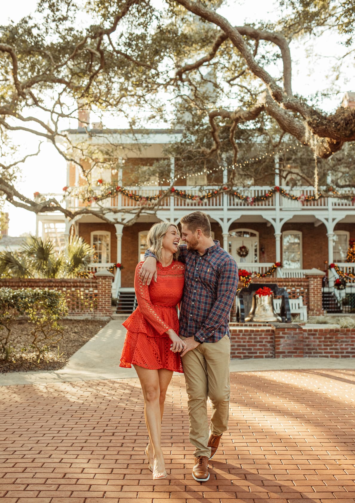 Couple walking arm in arm under live oaks in front of the holiday-decorated keeper's house