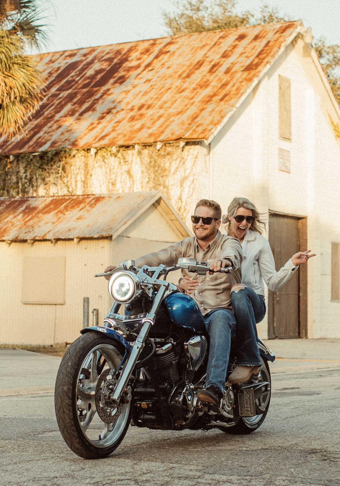 Laughing couple riding a blue motorcycle past a rusty tin-roof building in St. Augustine