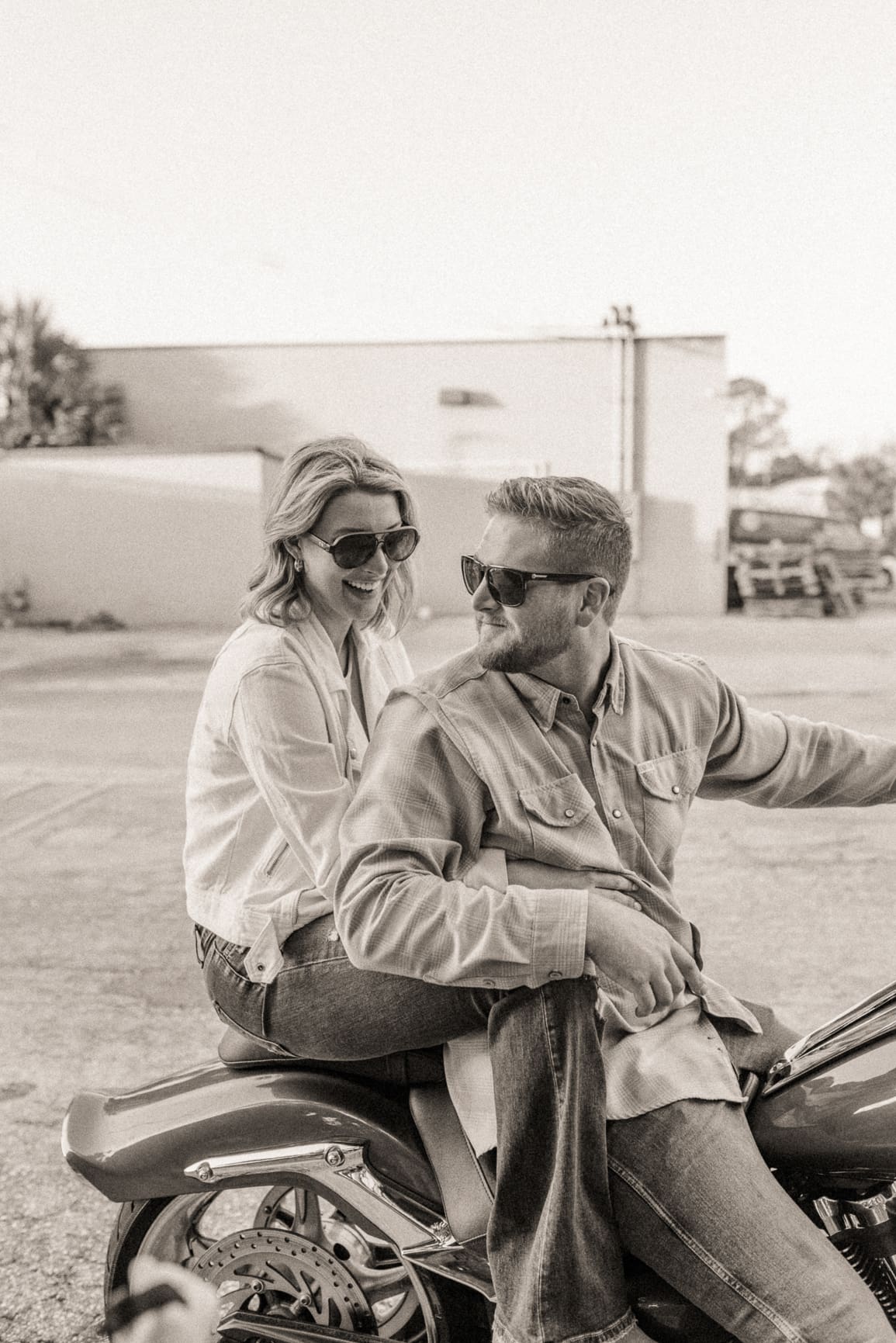 Black and white photo of an engaged couple in sunglasses smiling at each other on a motorcycle