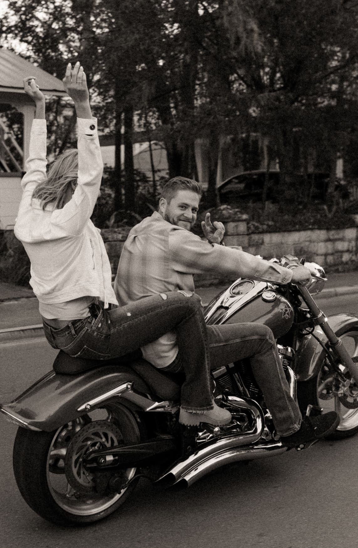 Black and white photo of a woman throwing her arms up on the back of a motorcycle as her fiancé smiles back
