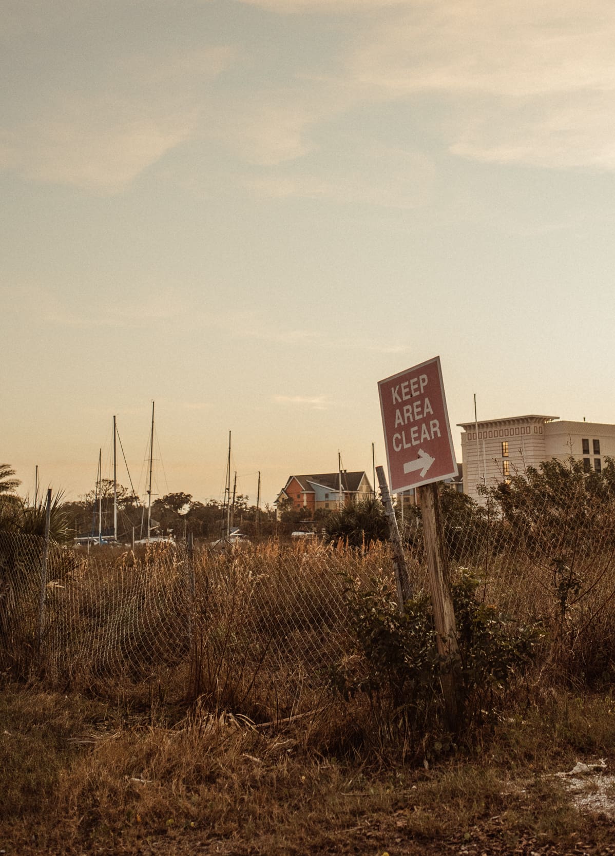Weathered "Keep Area Clear" sign in an overgrown lot, with sailboat masts behind it in St. Augustine