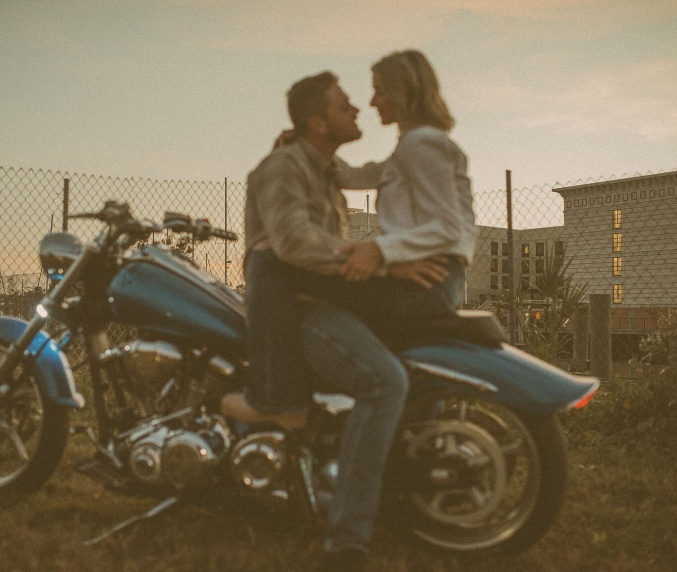 Couple leaning in for a kiss on a motorcycle beside a chain-link fence at dusk