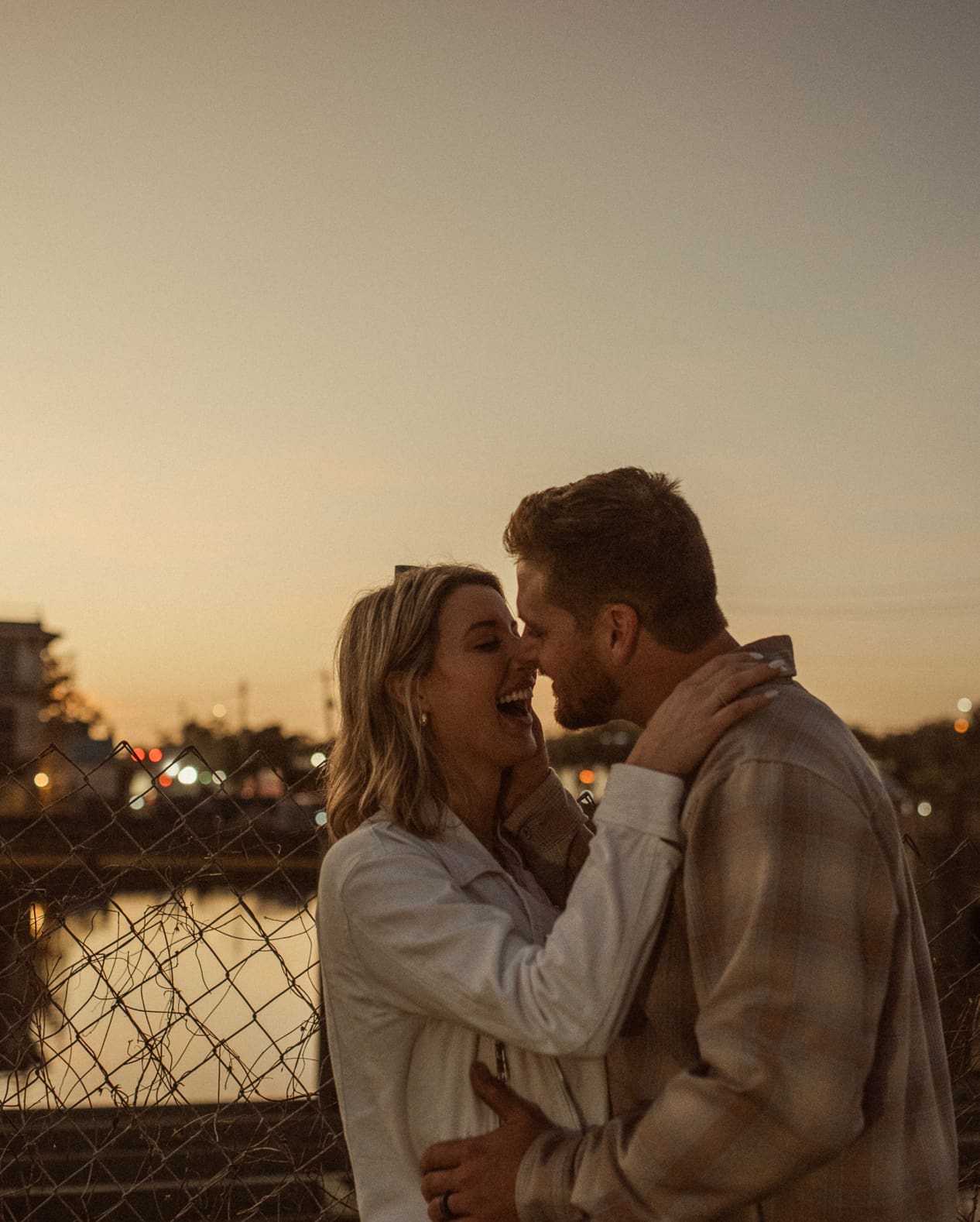 Couple laughing nose to nose beside a chain-link fence as city lights glow across the water
