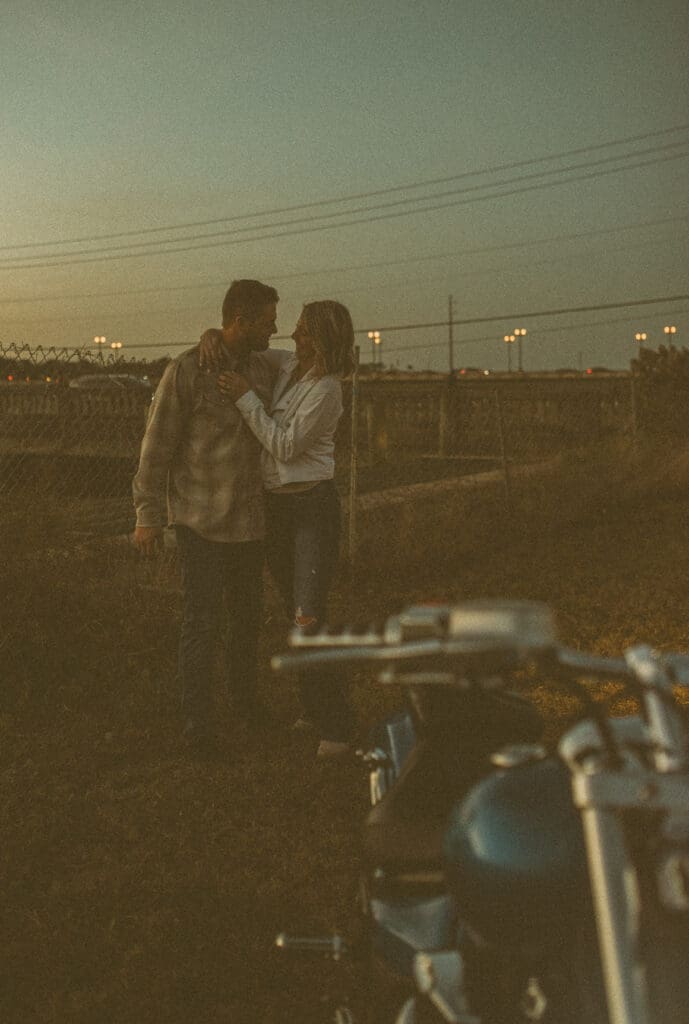  Couple standing close in an open field at dusk with the motorcycle in the foreground