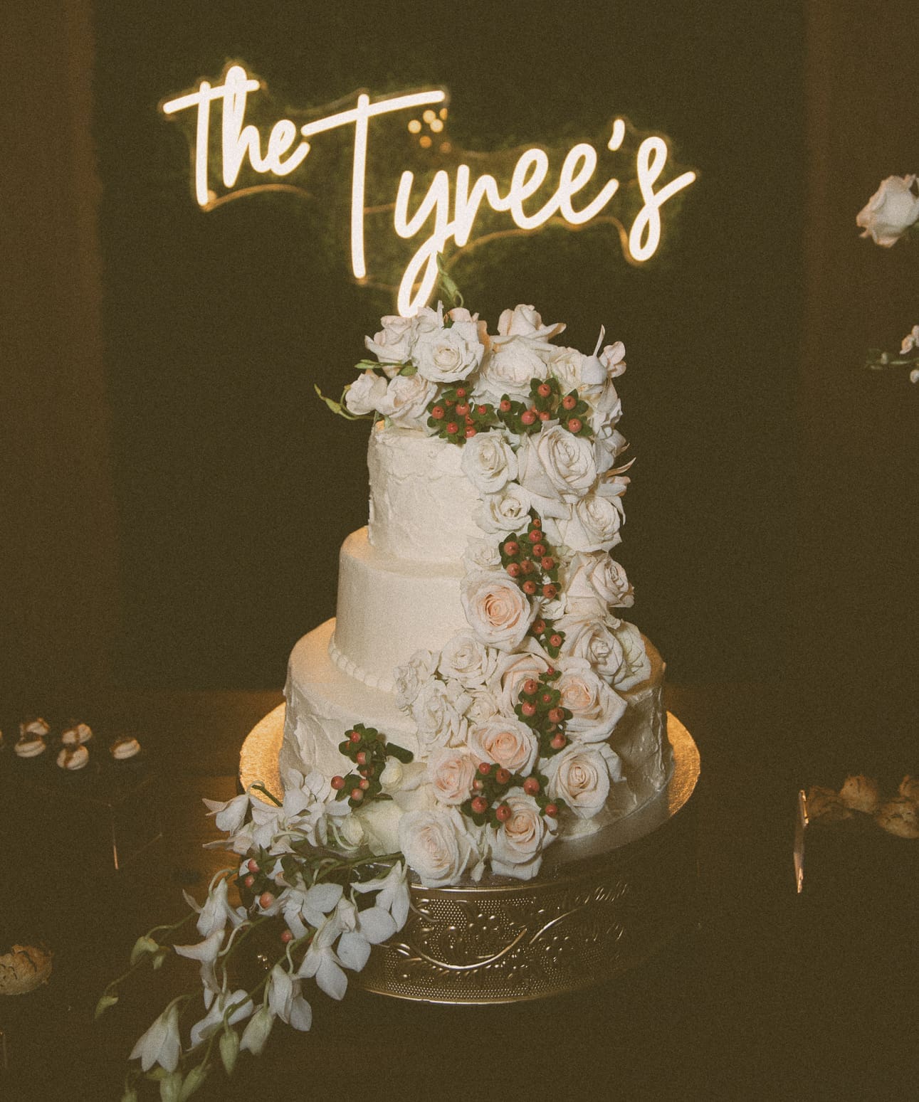 Three-tier wedding cake with cascading roses and a neon lit "The Tyrees" sign behind it. 