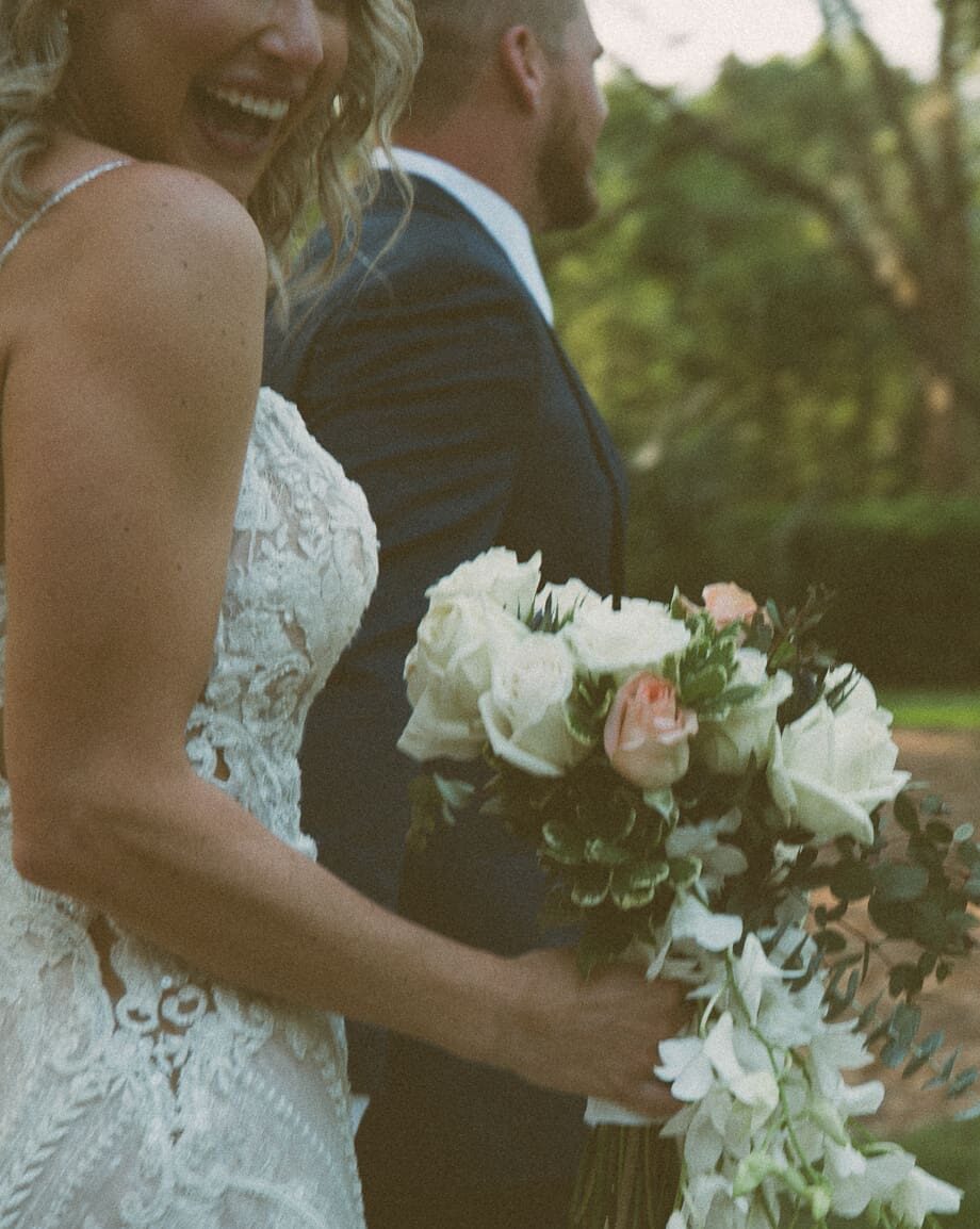 close-up of bride laughing with groom, white and blush bouquet in golden evening light at Bowing Oaks. 