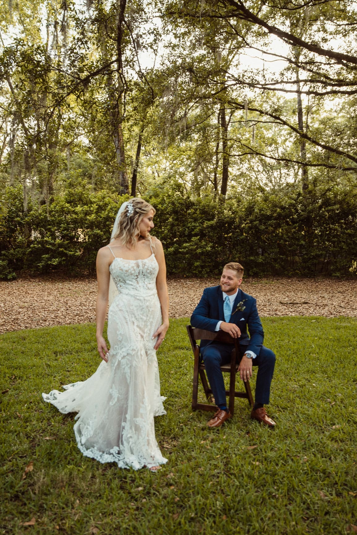 Bride standing beside seated groom in a wooded clearing at golden hour, Bowing Oaks wedding. 