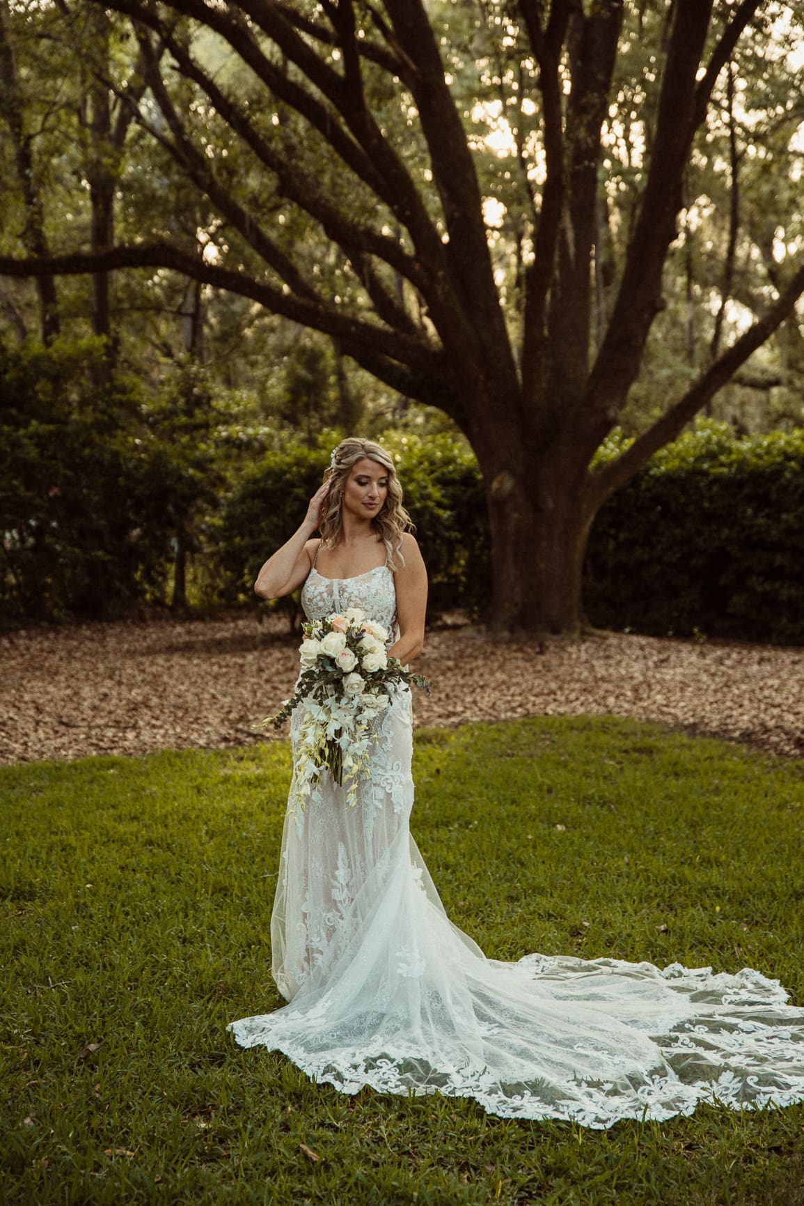 Full-length bridal portrait with lace gown and bouquet in front of a live oak at Bowing Oaks. 