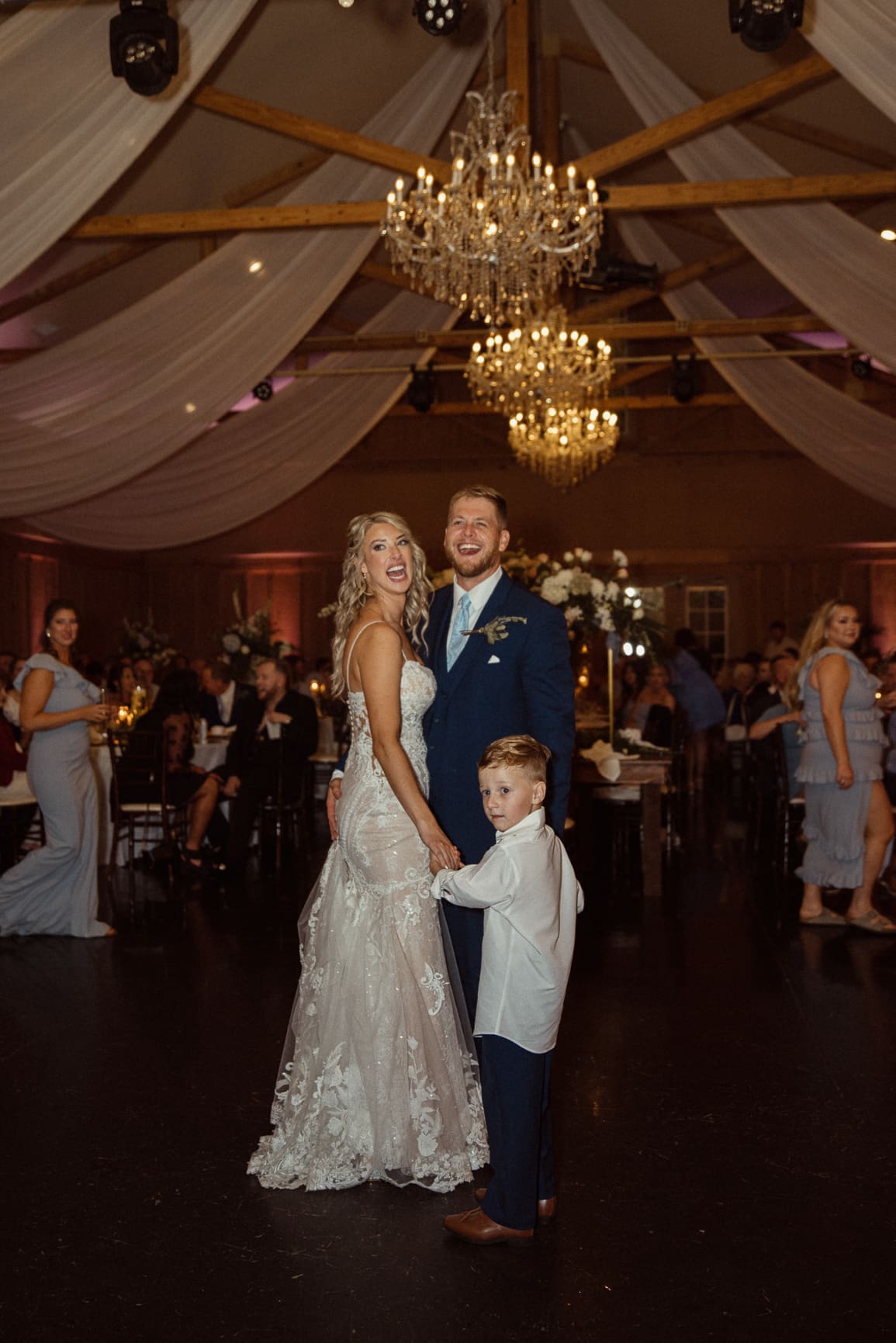 Bride and groom posing with young ring bearer beneath crystal chandeliers at Bowing Oaks reception. 