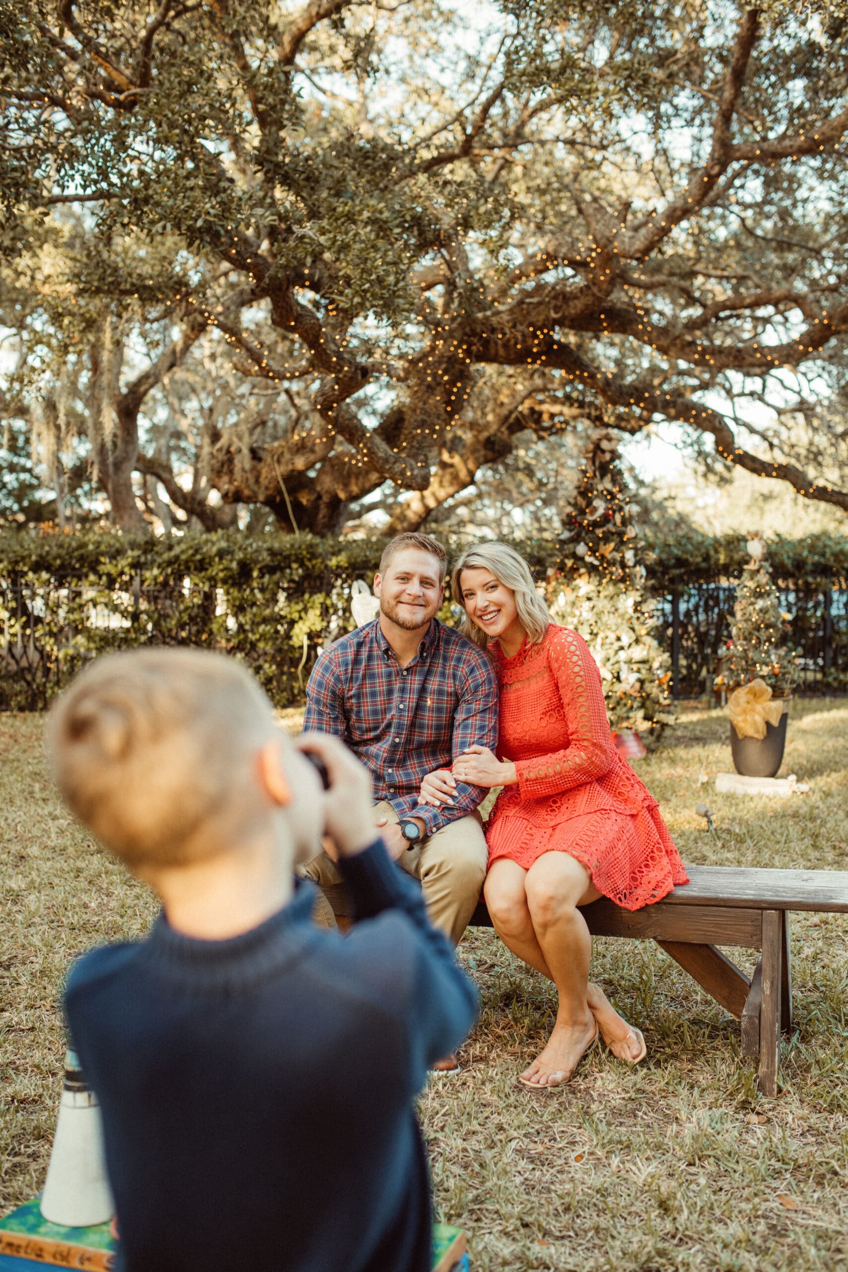 Young boy photographing his dad and future stepmom on a bench under string-lit live oaks