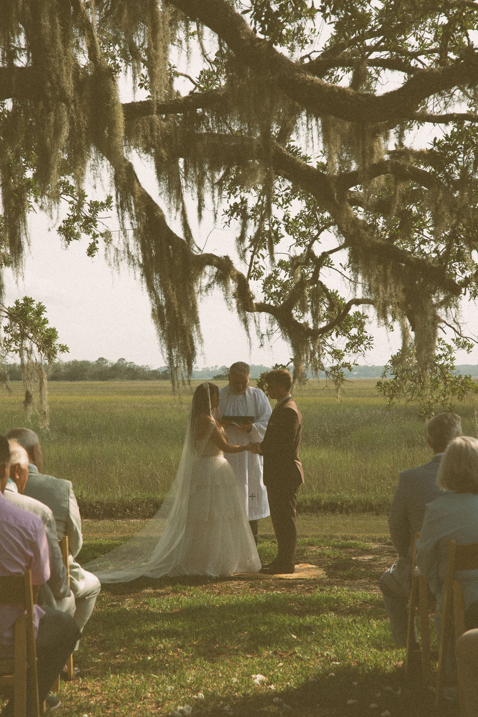 Couple holding hands during their ceremony beneath a moss-draped oak overlooking the marsh, shot on 35mm color film.