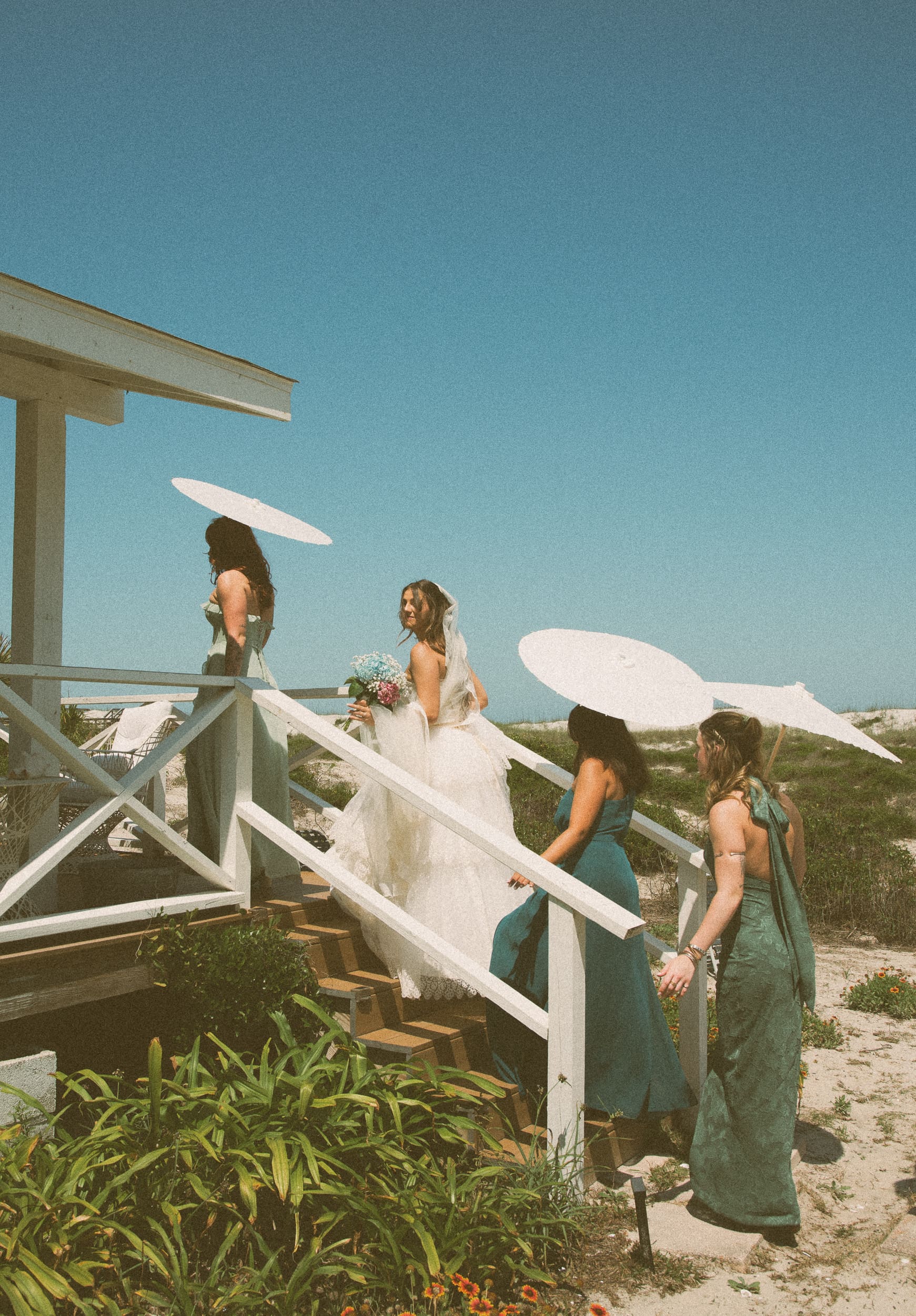 Bride and bridesmaids in green with white parasols climbing beach stairs under a blue sky, shot on color film.