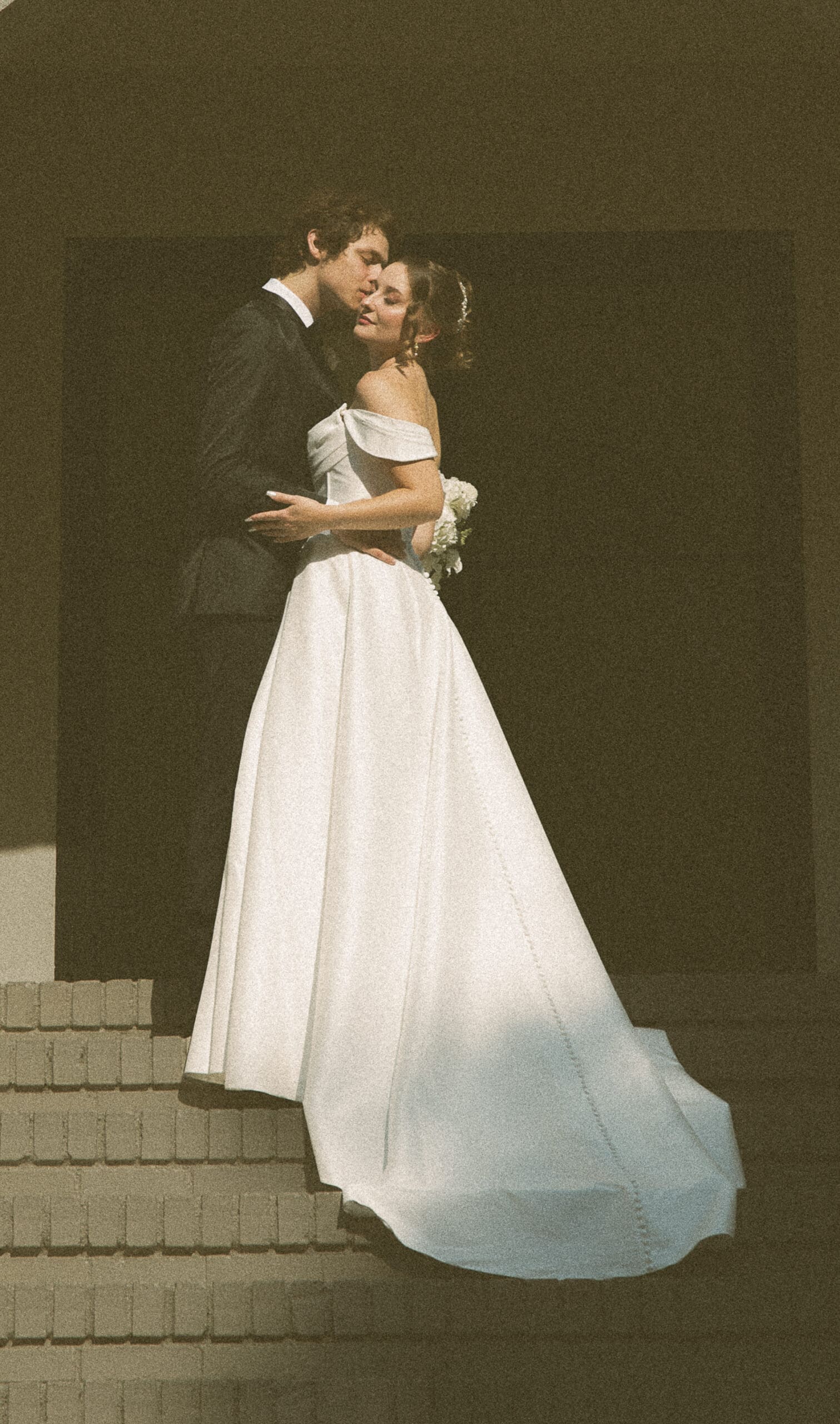 Groom kissing his bride's cheek as they embrace on brick steps, her off-the-shoulder gown catching the light, on 35mm film.