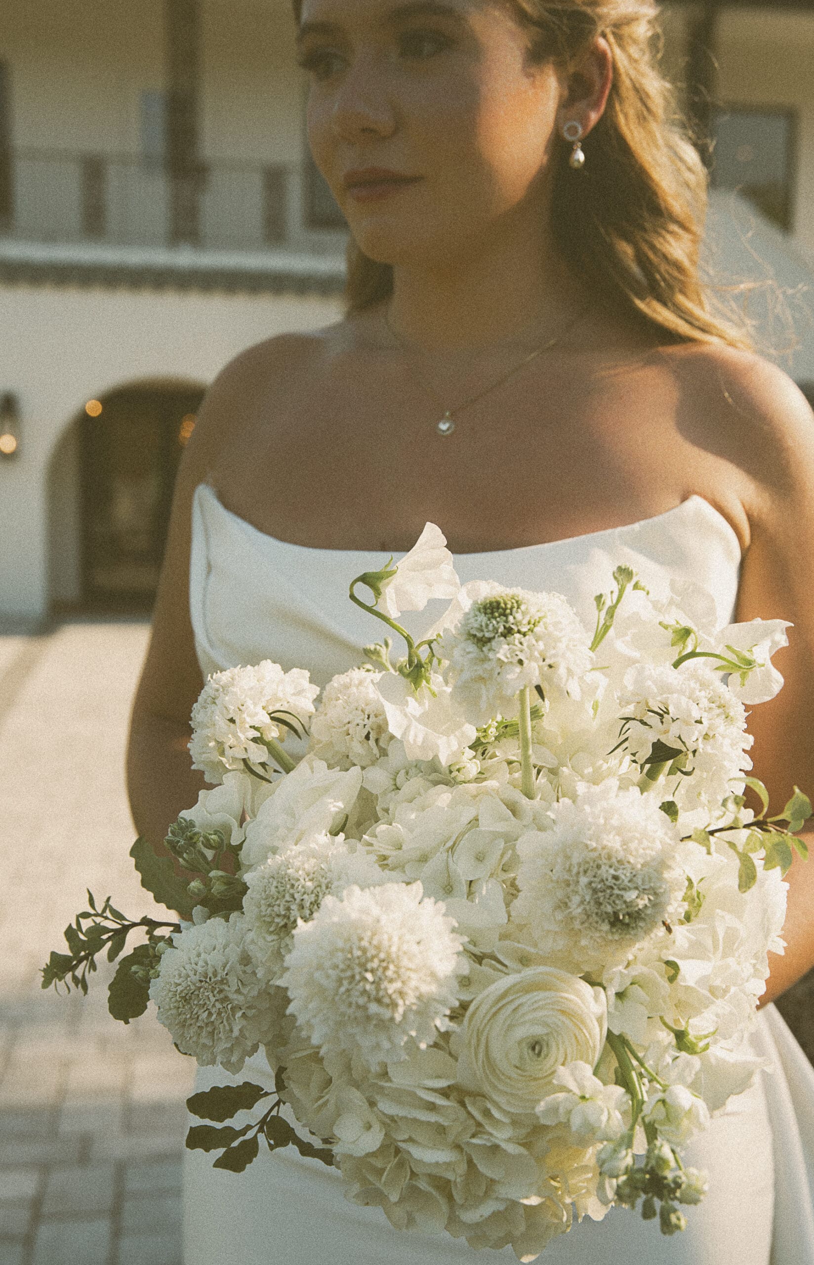 Bride holding a lush white bouquet in soft afternoon light in front of a Spanish-style building, shot on color film.