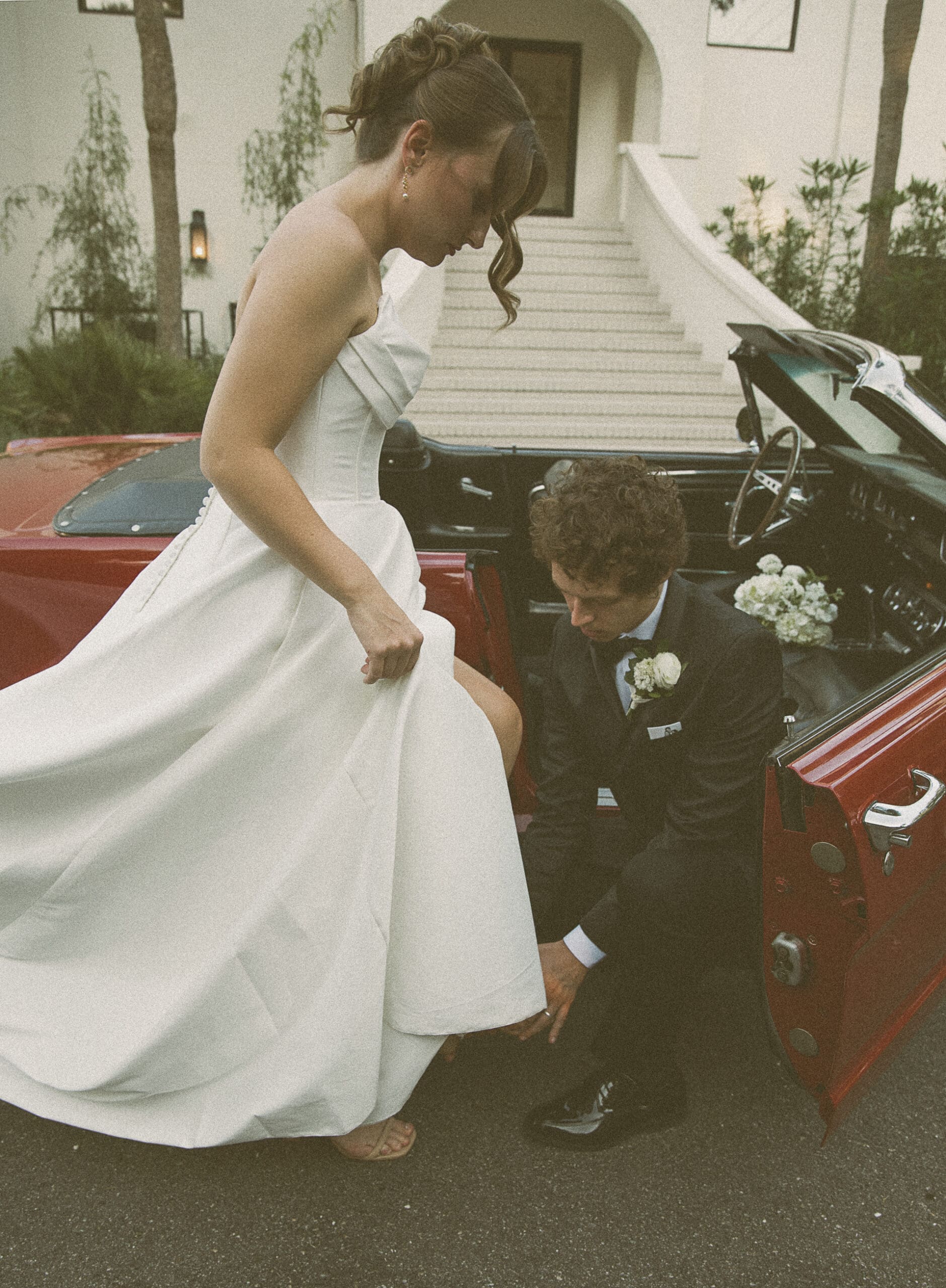 Groom kneeling to help his bride into a vintage red convertible in front of a Spanish-style building, shot on film.