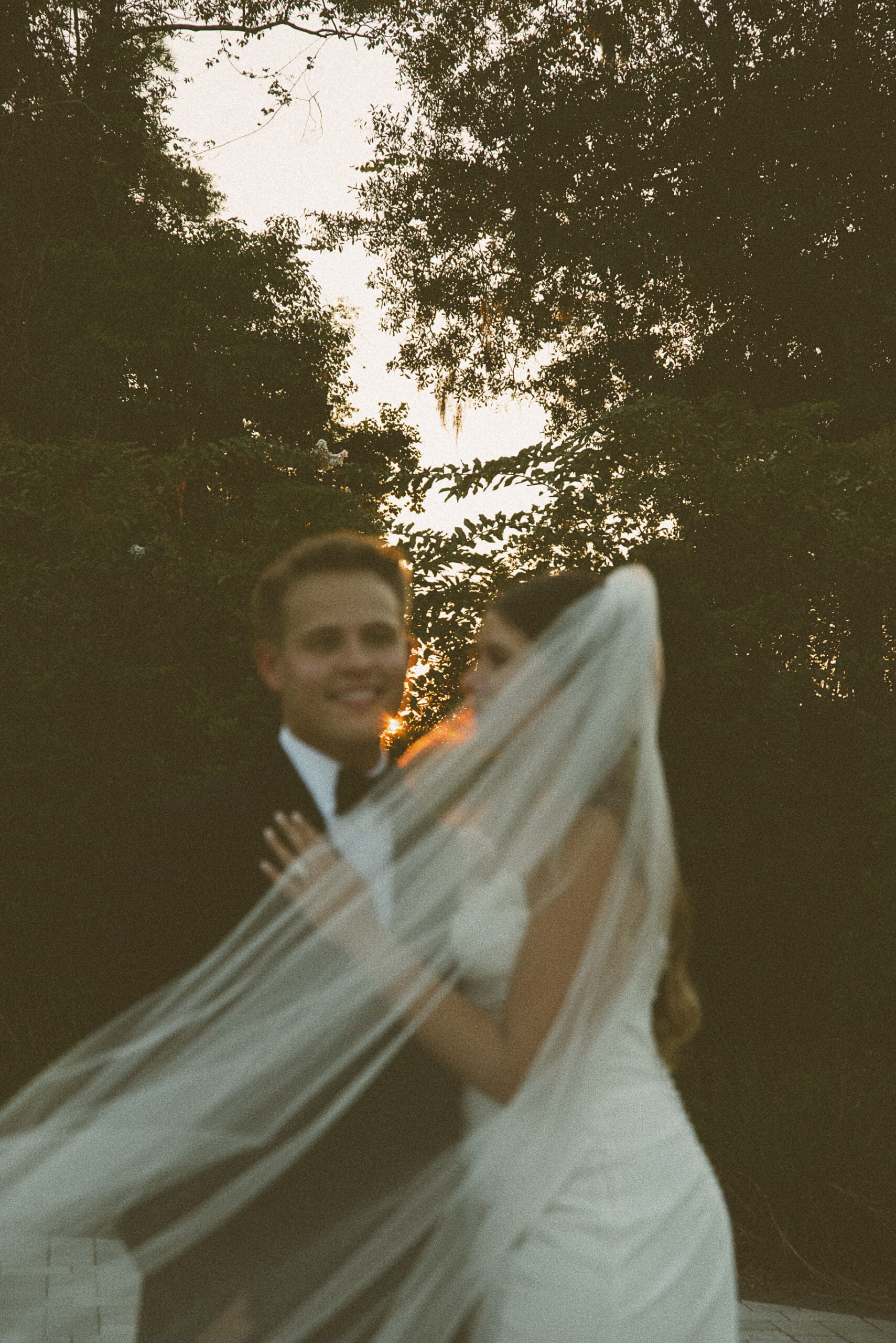Couple embracing at sunset beneath moss-draped trees as the bride's veil drifts across the frame, captured on film.