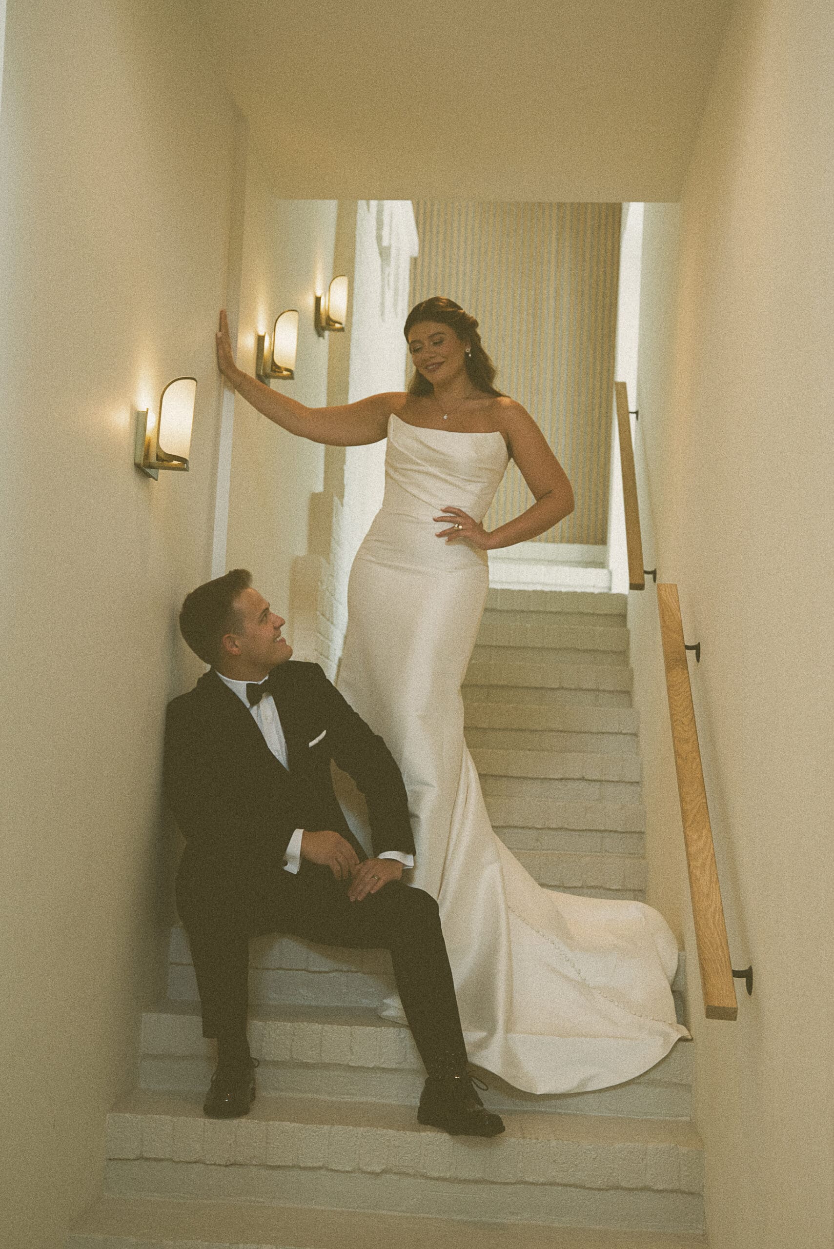 Bride leaning against a sconce-lit wall on a staircase as her groom sits below smiling up at her, on film.