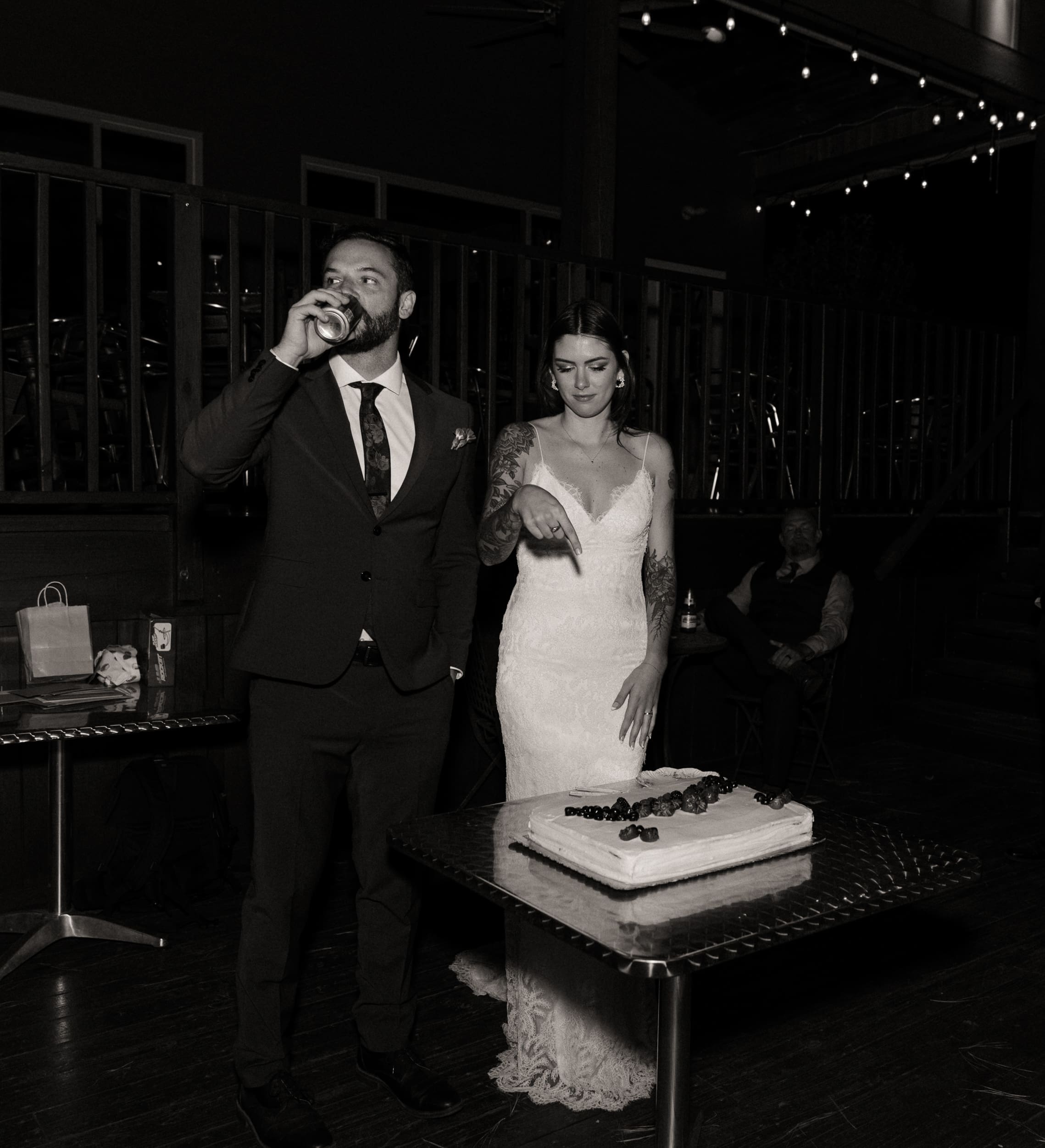 Groom taking a drink as his bride points to the cake during the reception, on black and white film.