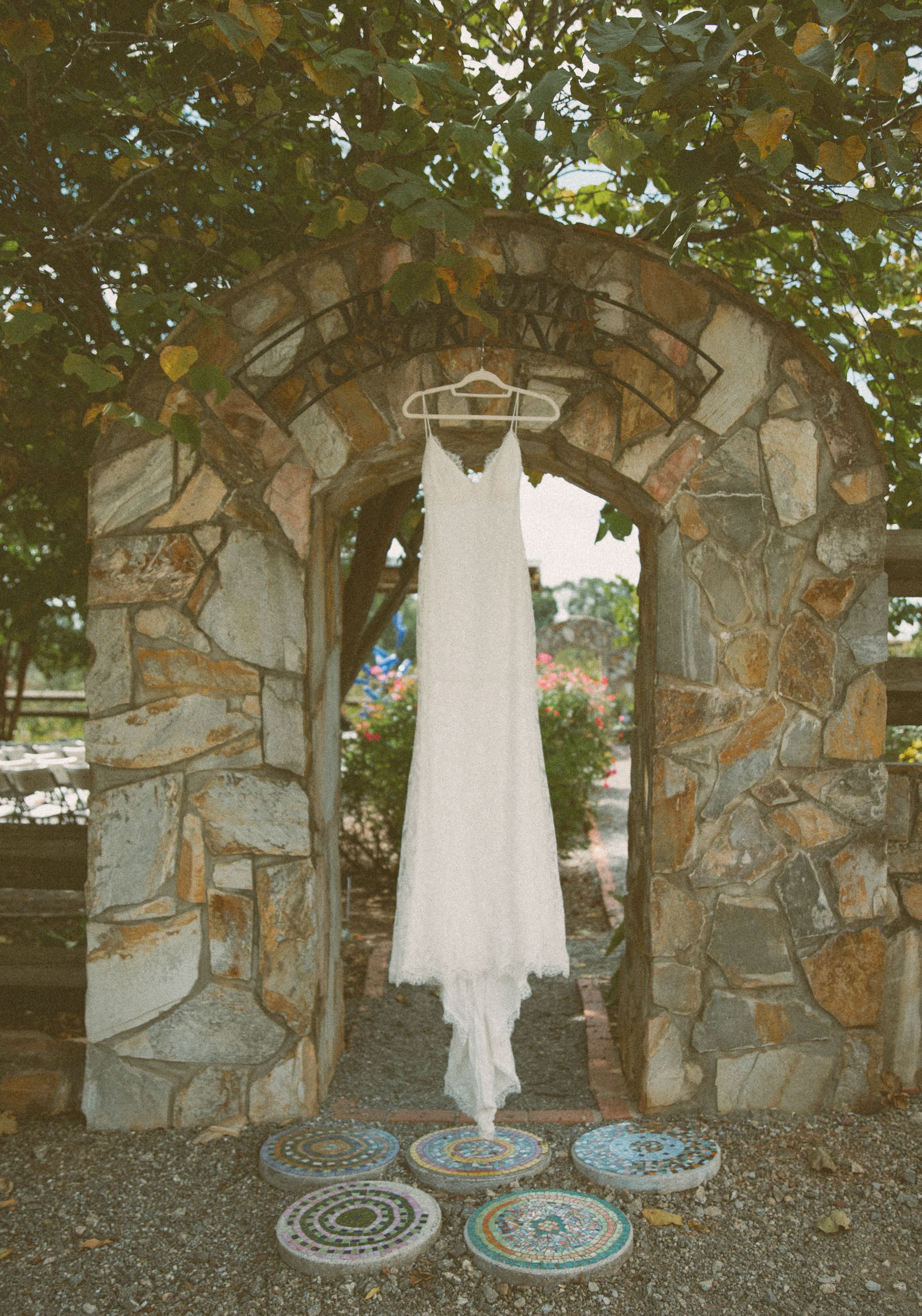 Wedding dress hanging in a stone archway beneath green leaves, photographed on film.