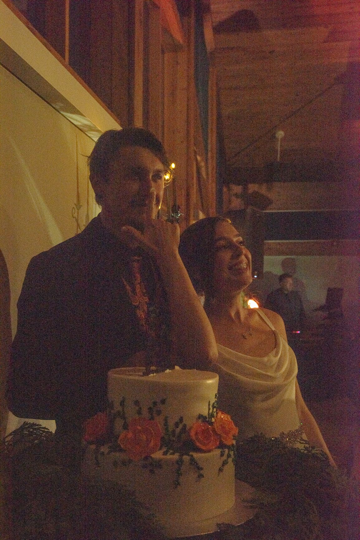 Groom tasting frosting as his bride smiles beside their rose-topped cake, glowing in warm reception light on film.