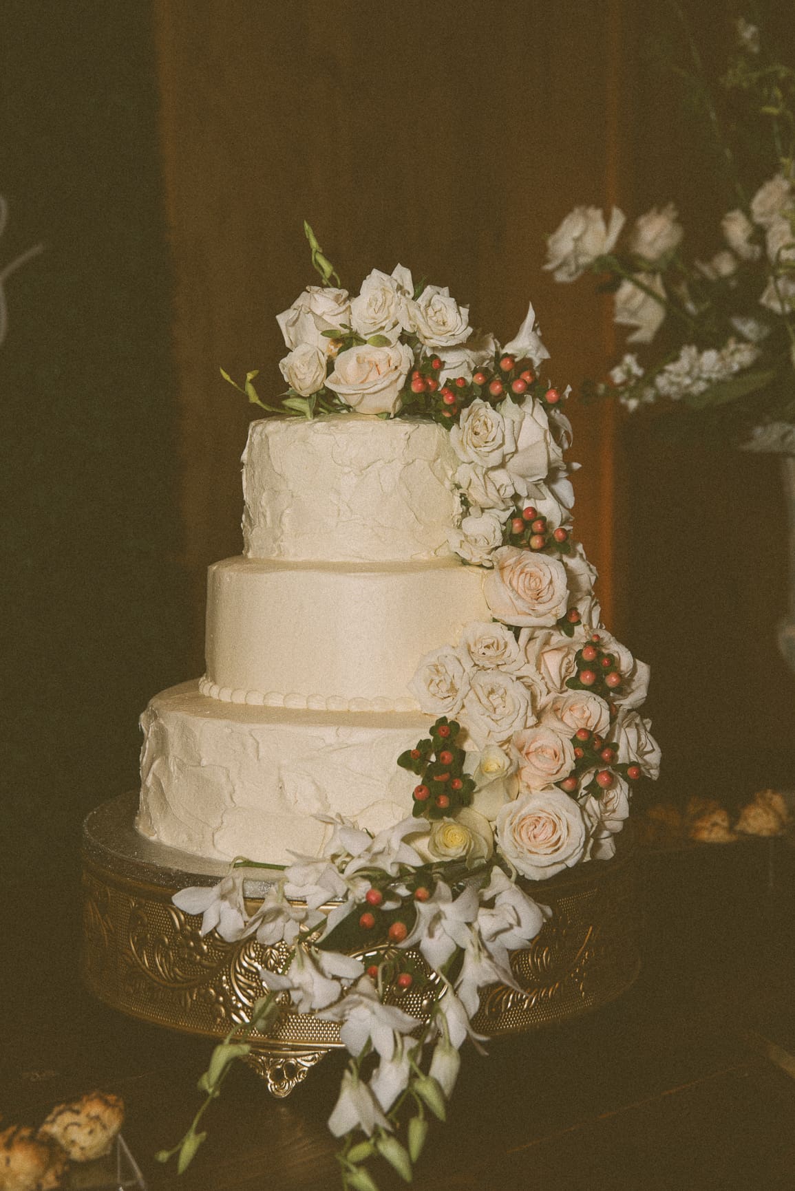 Three-tier wedding cake with cascading roses and berries, glowing in warm film tones.