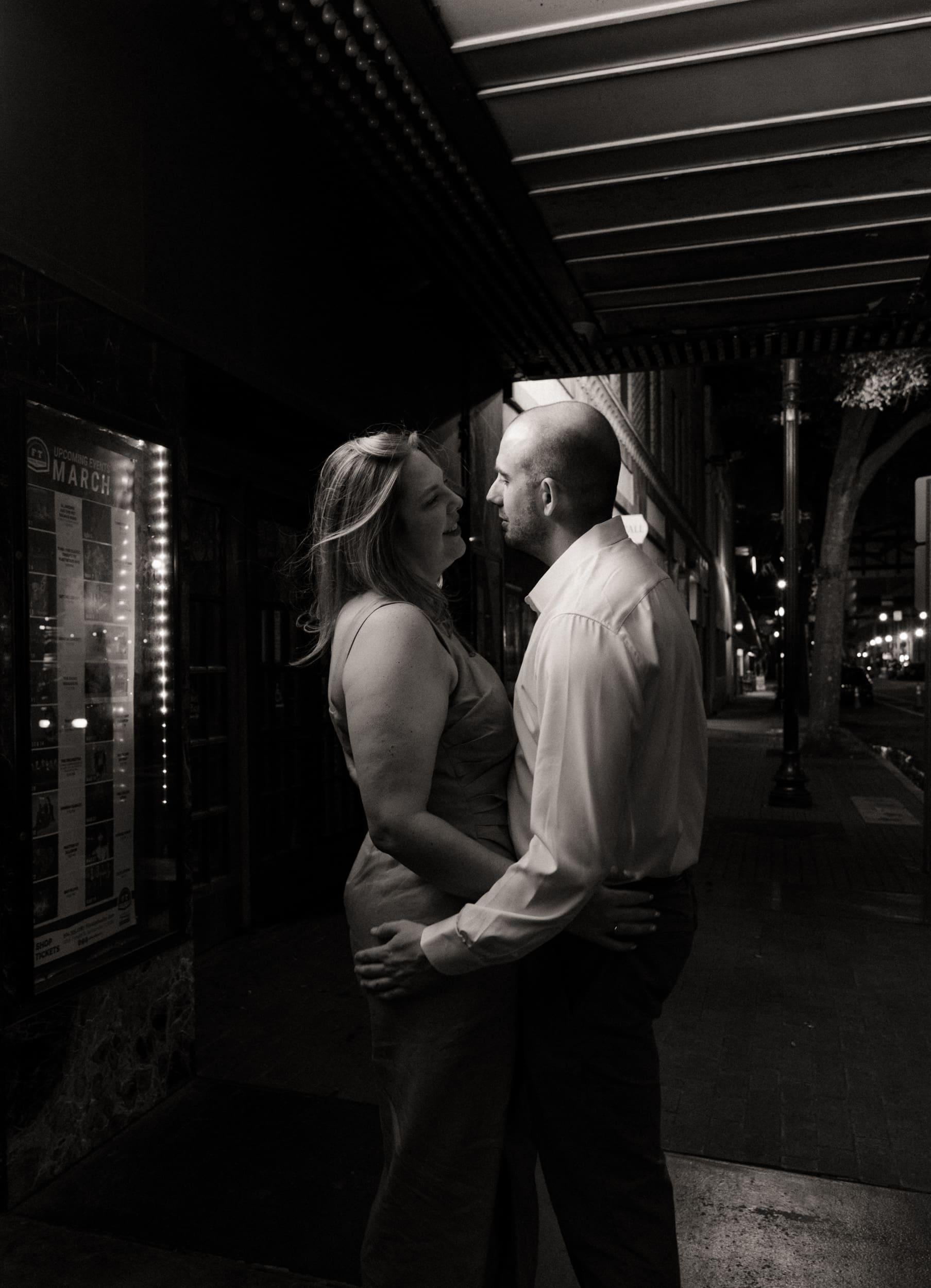 Black and white photo of an engaged couple face to face beneath a theater marquee at night.