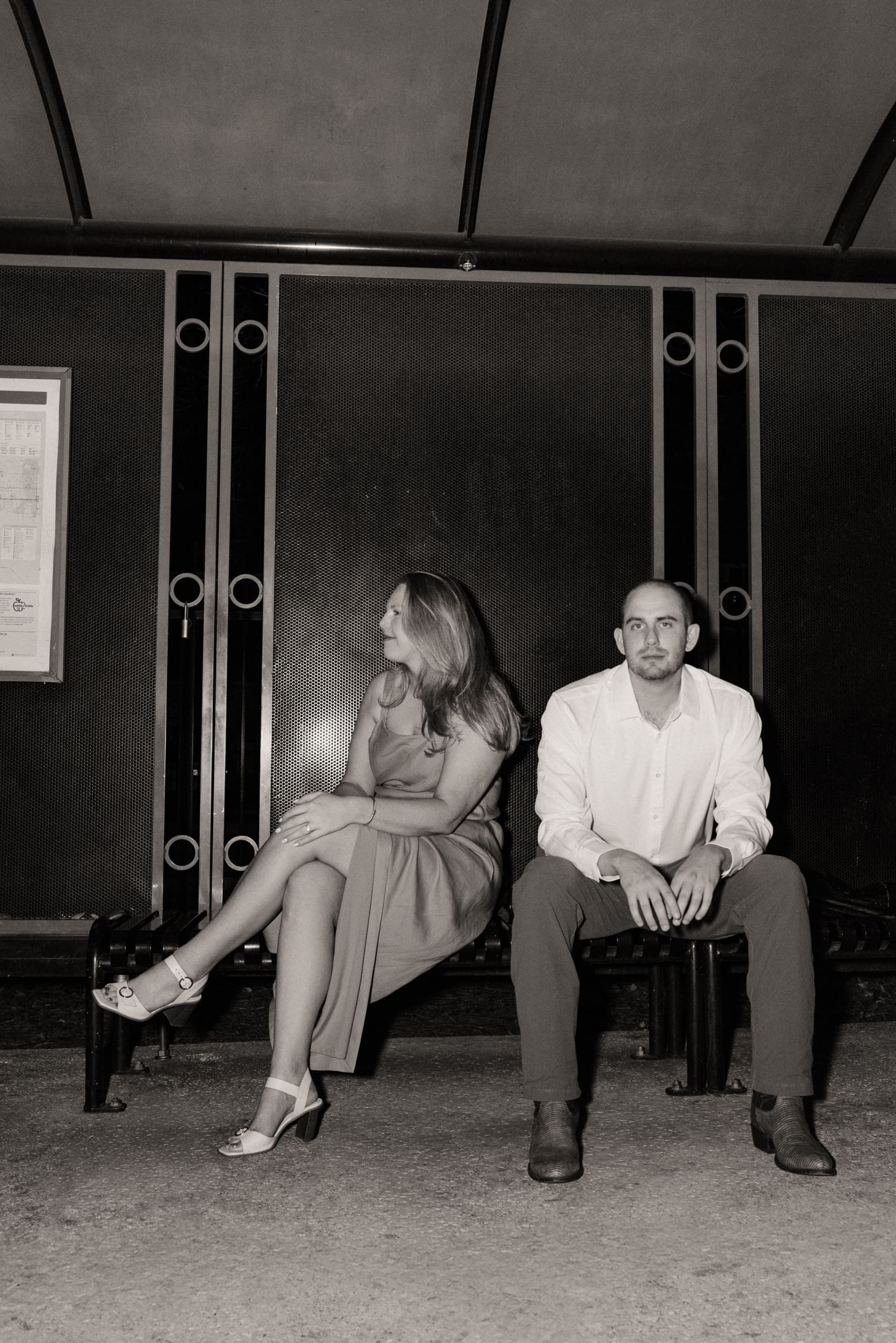 Black and white photo of a couple sitting on a downtown bus stop bench as she glances toward him