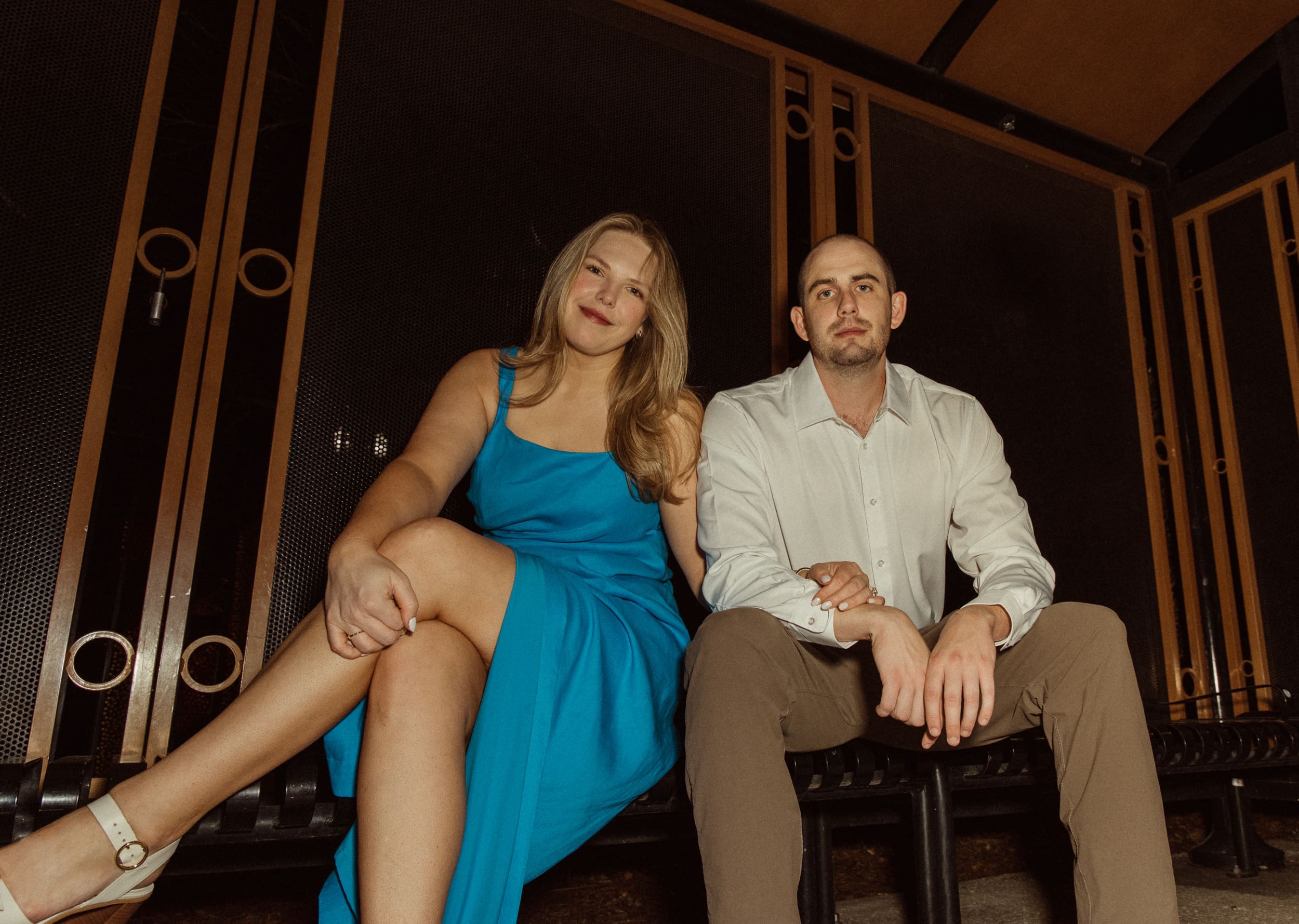 Woman in a blue dress holding her fiancé's arm as they sit at a bus stop at night
