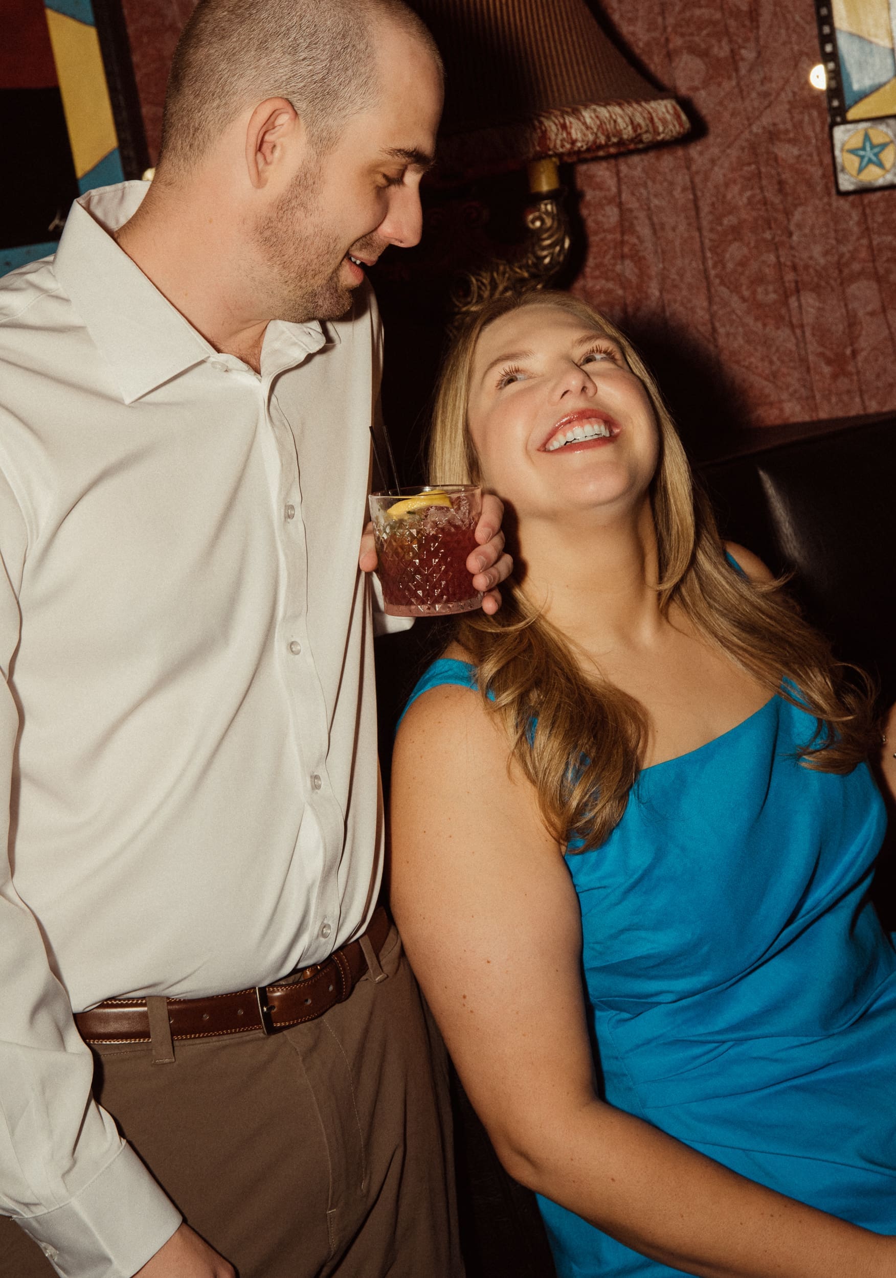 Woman in a blue dress laughing up at her fiancé as he holds a cocktail beside a vintage fringed lamp