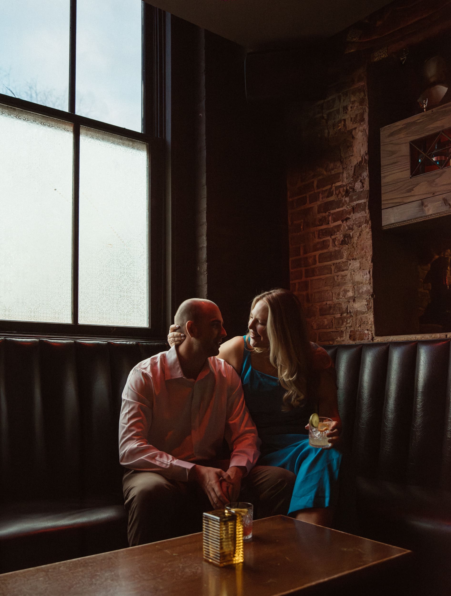 Woman cupping her fiancé's face in a leather booth by a frosted window and exposed brick at Dos Gatos