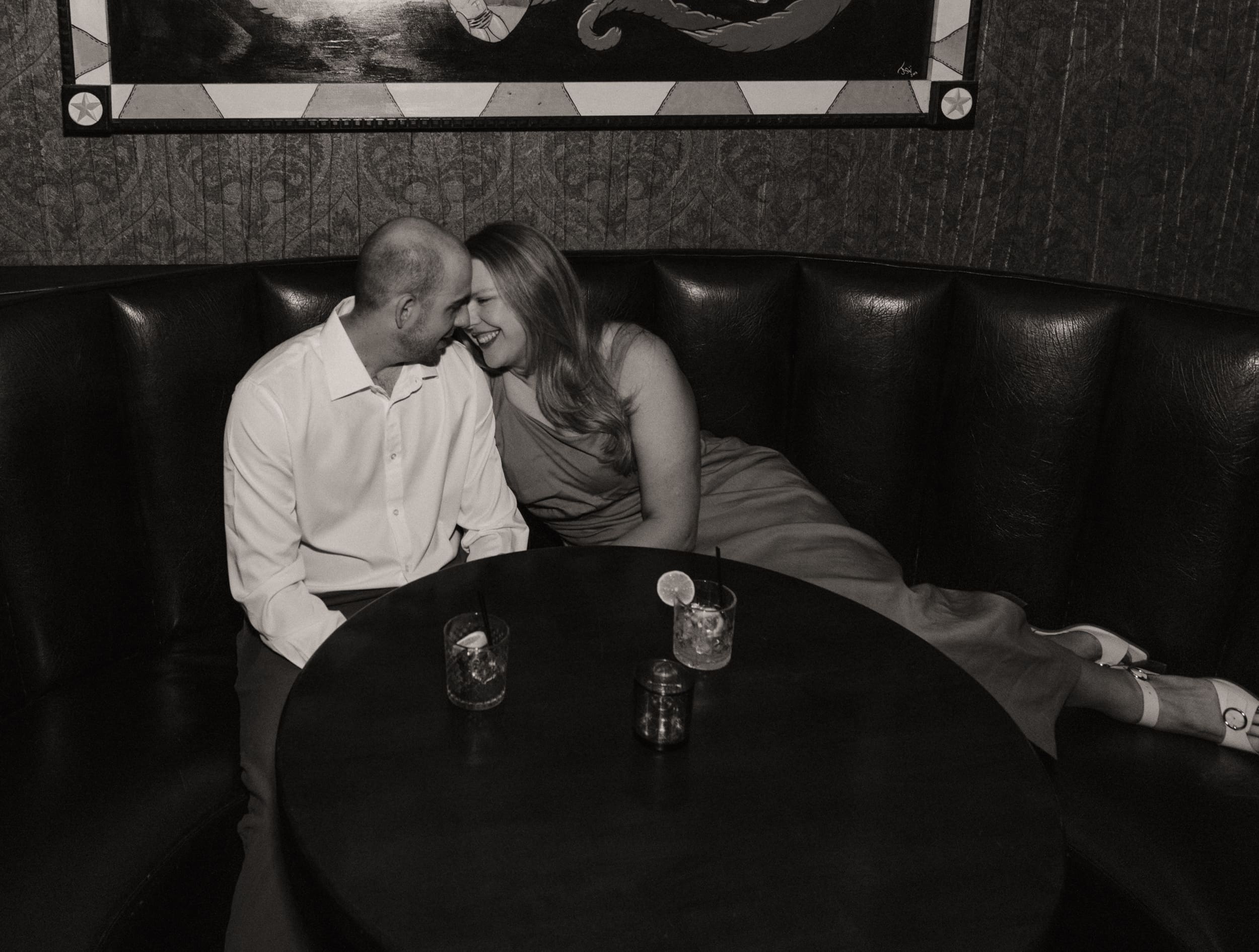 Black and white photo of an engaged couple touching foreheads and smiling in a leather booth